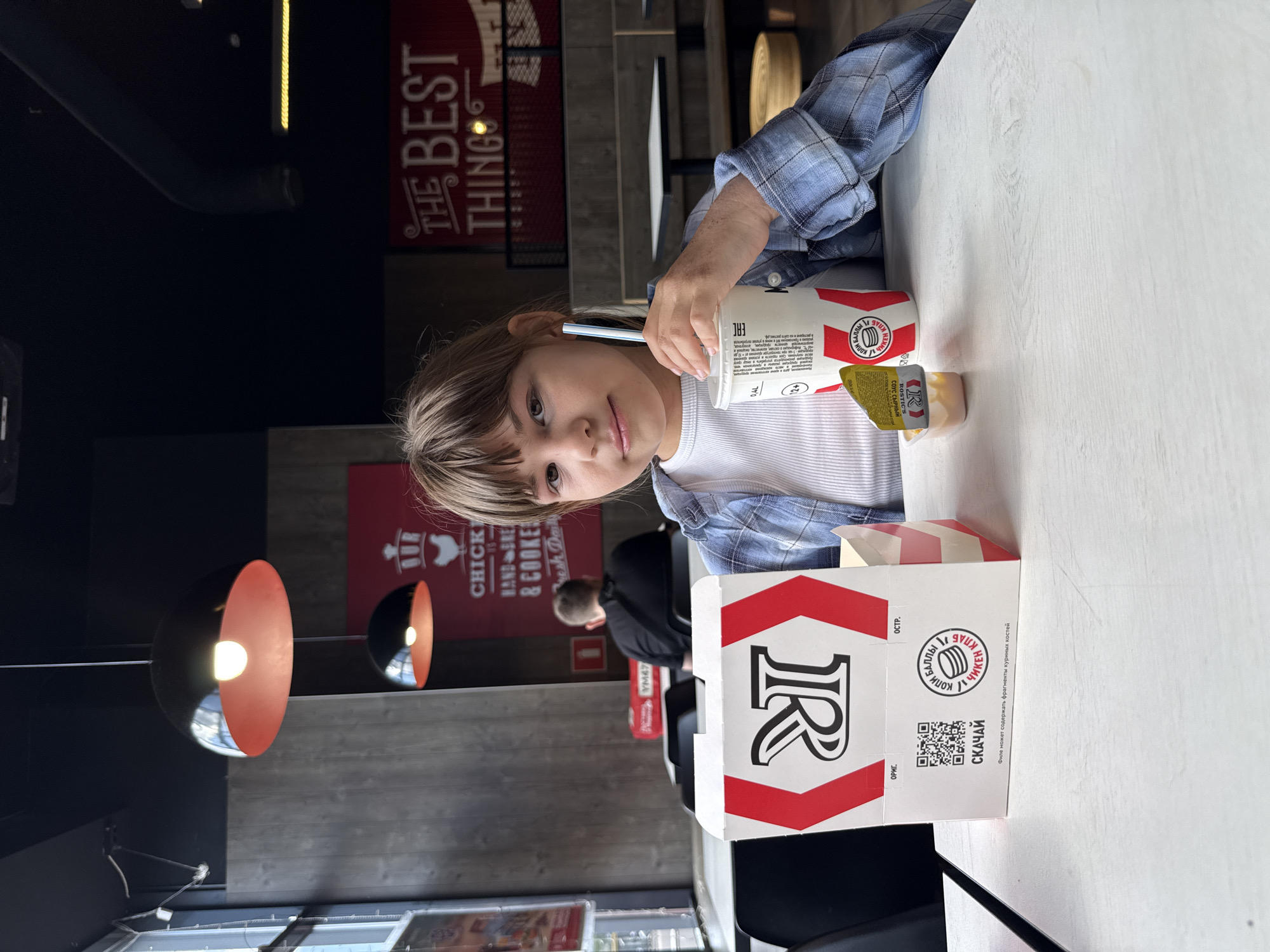 A child is sitting at a table in a restaurant, holding a straw in a cup. The restaurant appears to be a Rostik's based on the branding on the cup and box, and it is located in the city of Zavolzhye, Russia, as indicated by the Russian text on the box. The table is white and has a cup, a sauce container, and a food box with the Rostik's logo on it. The child is wearing a blue and white plaid shirt over a white shirt. In the background, there are other patrons and pendant lights with orange interiors. Signs related to the restaurant's menu are visible on the wall.