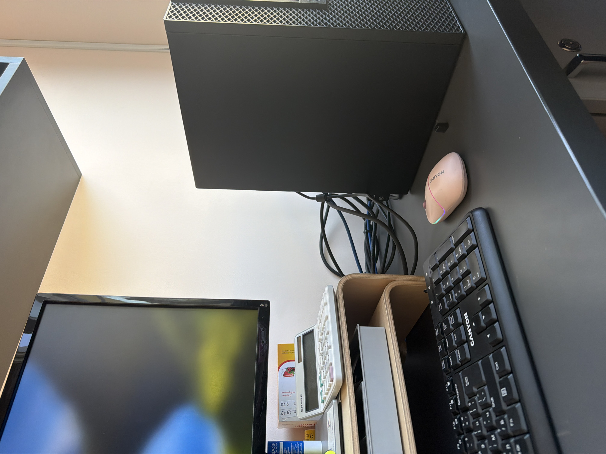 The image shows a desk setup in a room. On the desk is a black computer tower, a black keyboard with the brand "CANYON" visible, and a pink computer mouse with LED lights. To the left of the computer tower is a monitor displaying a blurred image. Below the monitor is a wooden monitor stand with a grey tray underneath. A white calculator with a pink and green keypad is placed on top of the monitor stand, next to some boxes. Cables are visible behind the computer tower. The walls appear to be painted a light color. The scene is well-lit, likely by natural light coming from the right side of the image. The image appears to be taken from a slightly elevated angle, looking down at the desk. There are no people or animals visible in the image. The overall environment suggests a home office or study space. The location is identified as Dolno Ablanovo, Bulgaria.
