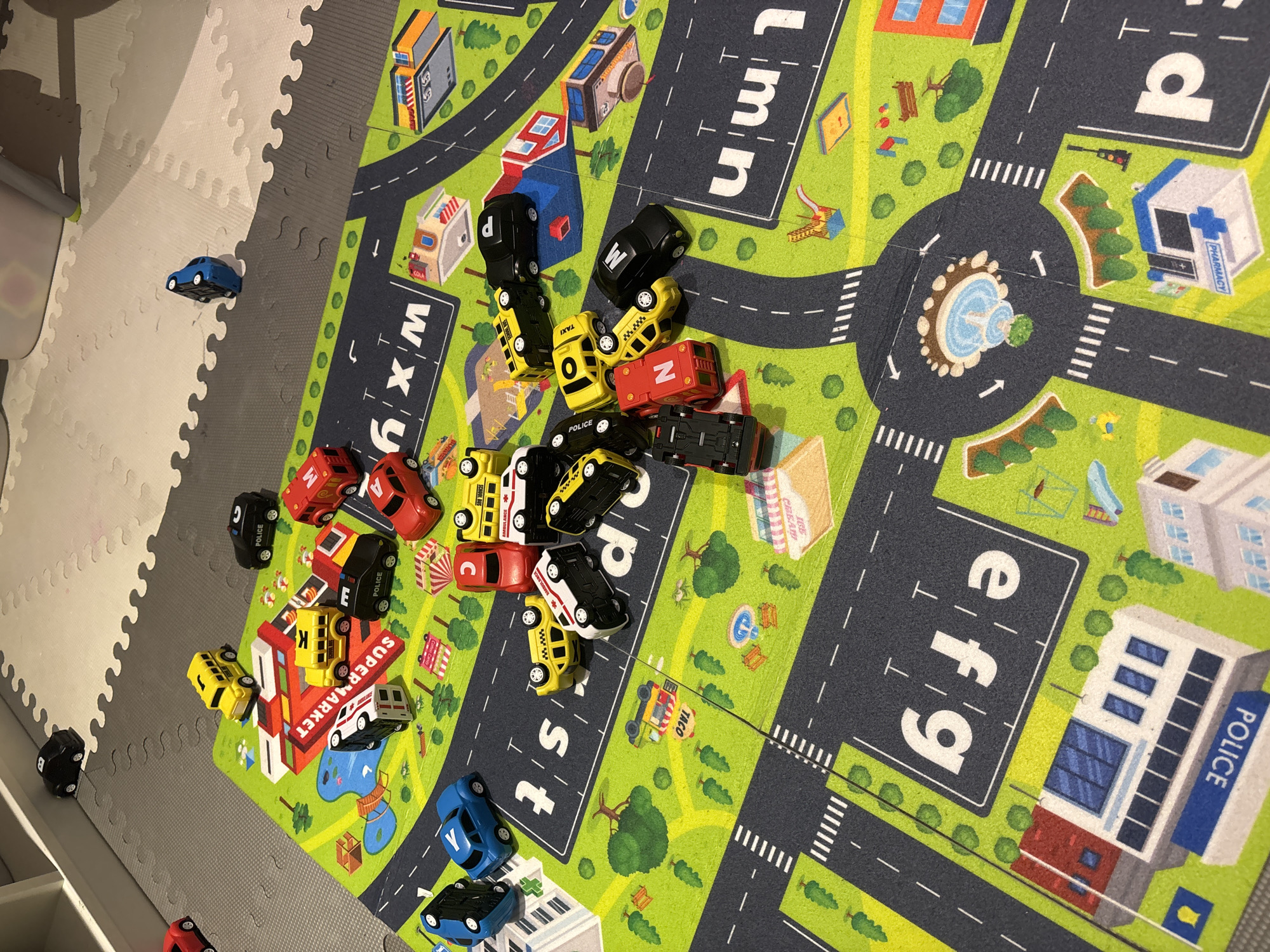 The image shows a toy car playset with various miniature vehicles scattered across a play mat depicting a cityscape. The play mat has roads, buildings labeled "POLICE" and "PHARMACY", a roundabout with a fountain, and green grassy areas with trees. The vehicles include police cars, taxis, ambulances, school buses, and what appear to be personal cars, in various colors such as black, yellow, red, and blue. Some cars are upright, while others are overturned or on their sides. The environment appears to be an indoor setting, with a portion of a white shelving unit visible in the upper left corner and a light-colored textured floor, possibly a foam mat, extending beyond the cityscape play mat. There are no people visible in the image, and no specific landmarks or indicators suggest the location is Dubai, United Arab Emirates. The scene is static, with no visible action or interaction between the toy vehicles.