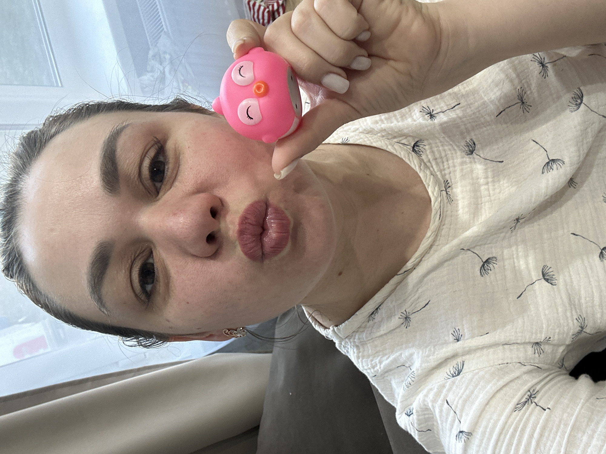 A selfie shows a woman holding a pink, owl-shaped object to her face. She is wearing a light-colored top with a dandelion-like pattern and has dark hair pulled back. The woman is pouting her lips. In the background, part of a window and a gray couch are visible. The location is Chaikovsky, Russia.