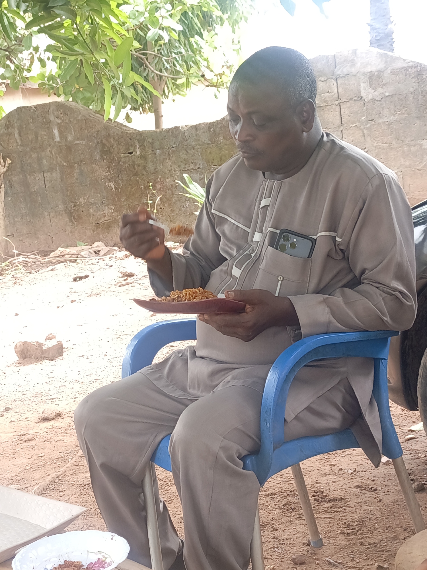 A man is seated outdoors in Akwanga, Nigeria, eating food from a brown plate with a fork. He is sitting in a blue plastic chair. The man is wearing a grey outfit with white trim and has a phone in his pocket. Behind him is a roughly constructed wall, and vegetation can be seen overhead. The ground is dirt and gravel. A white bowl with food remains is visible on a surface to his left.