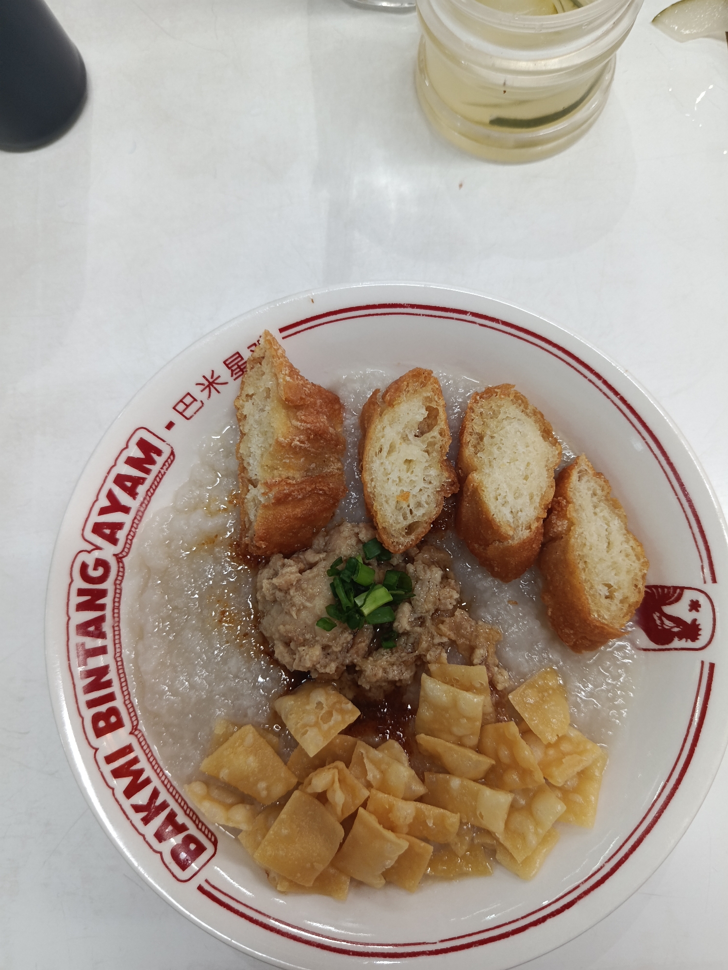 A close-up, overhead shot displays a bowl of congee topped with various savory ingredients. The congee itself is a creamy, pale white, with a slightly watery consistency, glistening with a sheen of oil. Scattered across the surface are crispy, golden-brown fried wonton wrappers, resembling small squares and rectangles. To the left of the wontons is a pile of minced meat, possibly chicken or pork, seasoned and slightly browned. Atop the minced meat are finely chopped green onions, adding a splash of color.
Dominating the upper portion of the congee are several pieces of fried dough, appearing golden-brown and airy, with a slightly irregular, crusty exterior. These pieces of fried dough are cut into roughly rectangular shapes.
The bowl itself is white with a red rim and red text encircling the outer edge. The text is in both English and what appears to be Chinese characters. The English text clearly reads "BAKMI AYAM" in a bold, stylized font. Above this, in smaller Chinese characters, it reads "巴米星".
The congee appears to be served as a meal, likely breakfast or a light lunch, given the texture and toppings. The lighting suggests an indoor setting, possibly a restaurant or home, with bright, even illumination. No people are visible in the frame. A glass with a pale yellow liquid and some green elements, possibly a drink, is partially visible in the top right corner. The overall impression is of a hearty and flavorful dish ready to be enjoyed.