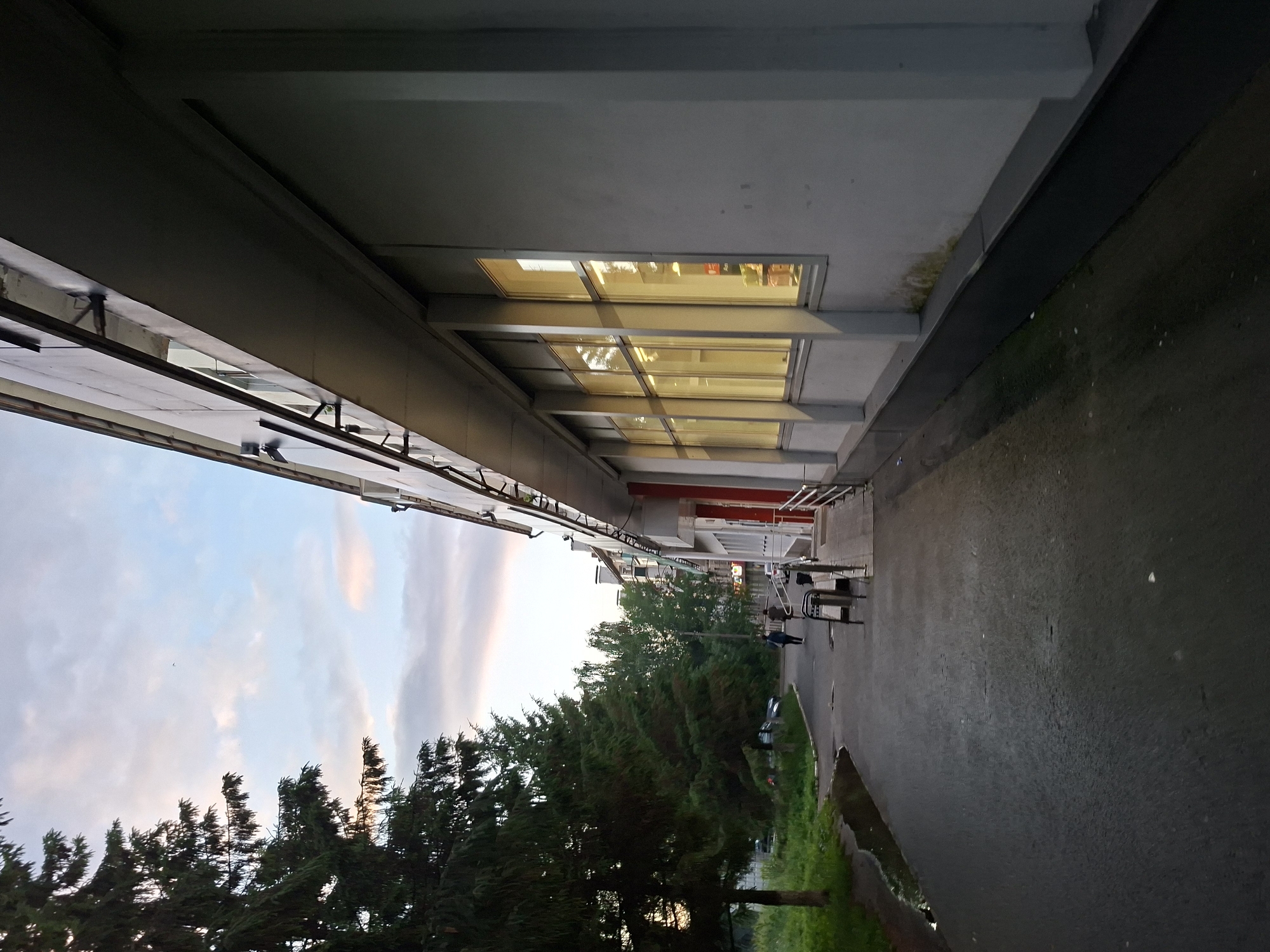 A dimly lit outdoor scene, likely an evening in a city, shows a paved walkway alongside a long, modern building with large, illuminated windows. Tall evergreen trees line the left side of the walkway, with a grassy area and some parked cars visible beyond. The sky is a muted blue with soft, colored clouds. Two individuals are walking away from the camera on the walkway, appearing to be moving towards the building or further down the path. The building's facade is light gray, and the walkway is dark asphalt, wet in patches suggesting recent rain. A metal trash receptacle and a ramp are visible near the building's entrance. The overall impression is one of urban infrastructure during twilight.