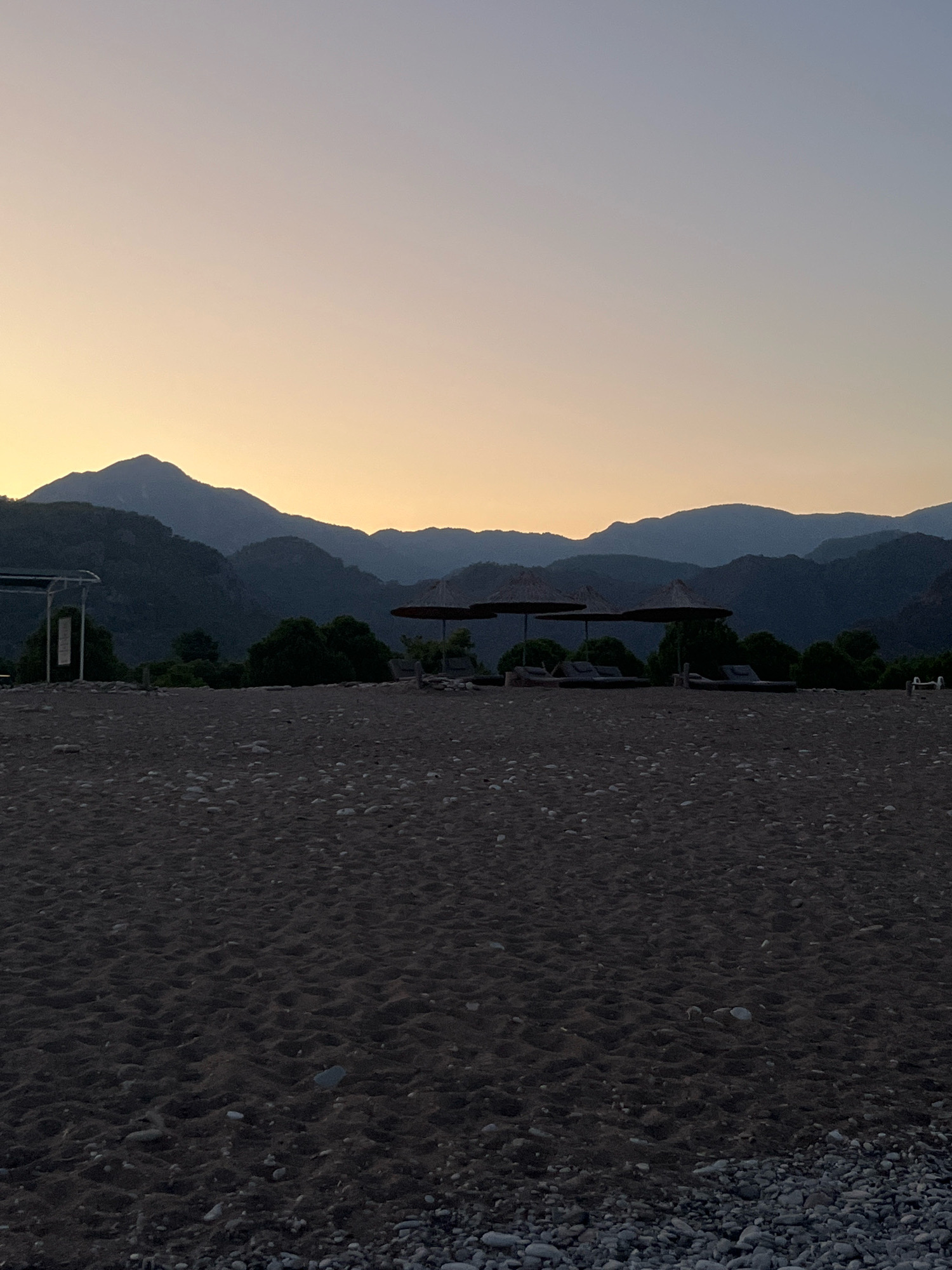 The image shows a beach scene at dusk in Kemer, Turkey. In the foreground is a dark, pebbly sand beach. In the middle ground, there are several beach loungers with thatched umbrellas, and some trees. In the background, silhouetted mountains rise against a pale orange and blue sky. The scene is tranquil and devoid of people.
