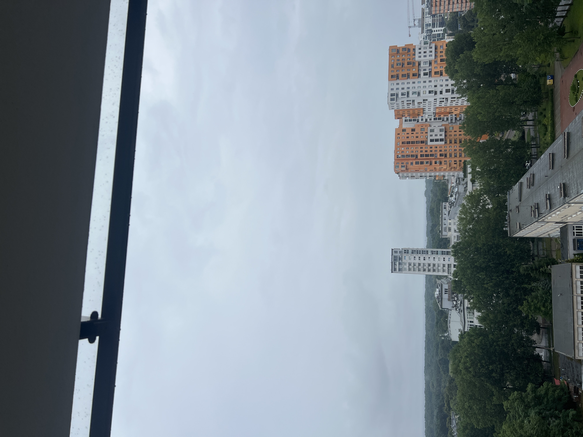 This is a wide-angle, eye-level exterior shot of a city skyline on a cloudy day, possibly with light rain. The foreground is dominated by a dark, horizontal structure that appears to be part of a balcony or window frame, with small water droplets visible on its surface and edge.
Below this frame, the scene opens up to a cityscape. In the mid-ground, several modern apartment buildings stand out. To the left, a tall, slender white building with numerous windows is prominent. To the right, a cluster of taller, more complex buildings with orange and white facades rise above the surrounding greenery. These buildings have a modular, multi-story design with many windows and balconies.
A dense line of lush green trees forms a natural barrier across the mid-ground, separating the buildings from a flatter landscape in the distance. Beyond the trees, the horizon is hazy and indistinct, suggesting a vast expanse of land or water, possibly a forest or park area, fading into the overcast sky.
The sky is uniformly gray and cloudy, with no visible sun or distinct cloud formations. The overall atmosphere is subdued and muted, consistent with overcast weather conditions. The image captures a view from an elevated position, looking out over the urban environment.
Based on the architectural style of the distinctive orange and white buildings, which are known as the "Oliwa Park" residential complex, this image is likely taken in the Oliwa district of Gdańsk, Poland.