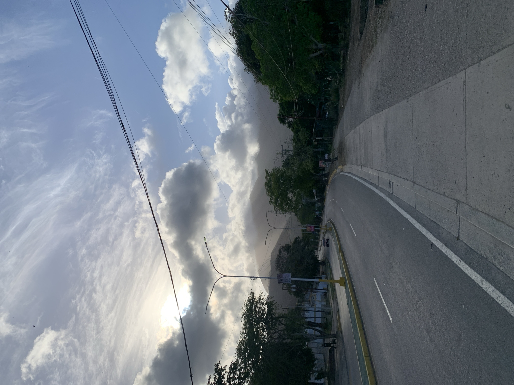 An eye-level shot captures a road in El Valle del Espíritu Santo, Venezuela, curving to the left as it recedes into the distance. The road is paved and marked with white lines. A concrete sidewalk runs along the right side of the road. On the left side of the road, there is a yellow-striped median and then another lane of traffic. Trees line the left side of the road, and beyond them, buildings are visible. A person is walking a dog on the left side of the road. In the distance, a car is driving on the road.
The sky above is partly cloudy with the sun peeking through some of the clouds, creating a hazy effect. Power lines are strung across the sky, supported by utility poles that are visible along the road. In the background, mountains are shrouded in mist and clouds. The overall impression is one of a somewhat overcast but scenic day in a town located near mountains.