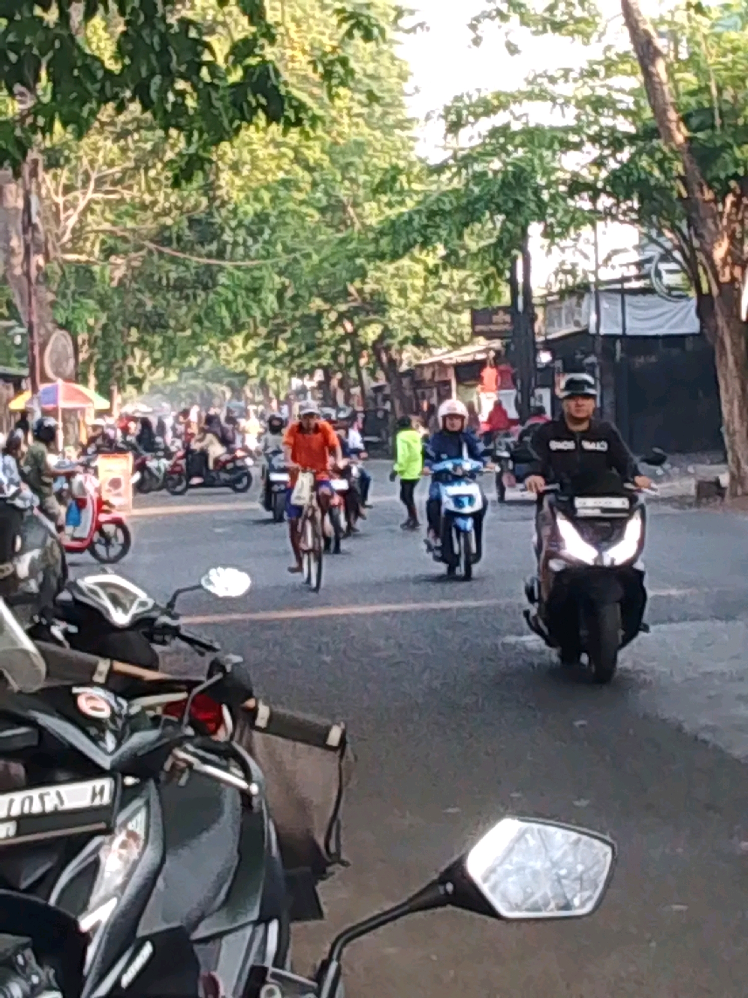 The image depicts a street scene in Probolinggo, Indonesia, likely during daytime. The street is lined with trees on both sides, casting shadows. Several people are riding motorcycles and a bicycle. In the foreground, a black scooter is parked. Further down the street, a person in an orange shirt is riding a bicycle. To the right, a person on a scooter wearing a black jacket and helmet is visible. On the left side of the street, there are some market stalls and shops. The overall atmosphere suggests a busy, urban environment with ongoing traffic.