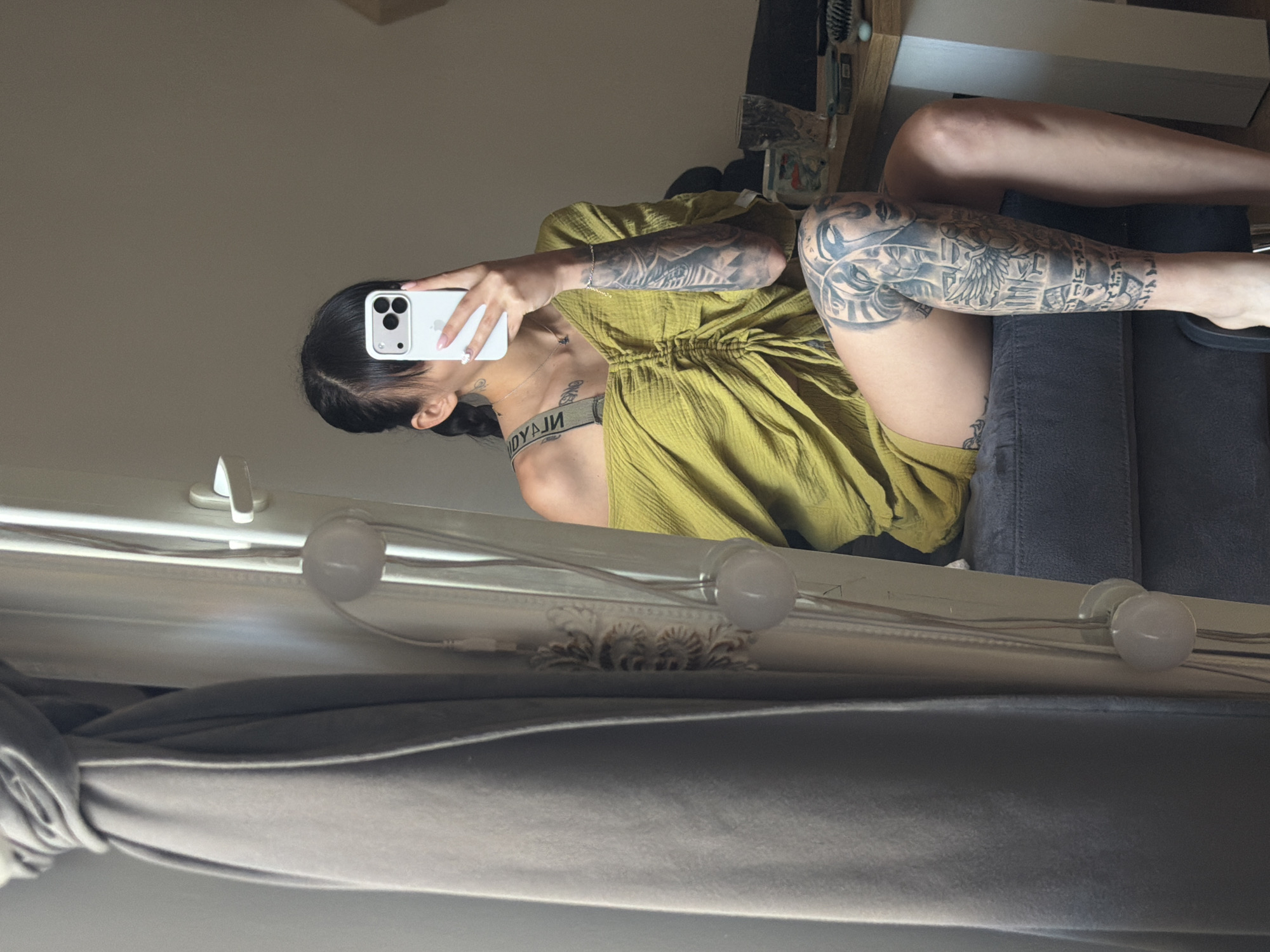 A woman with dark hair in a ponytail, wearing an off-the-shoulder yellow ruched dress, is sitting on a dark grey couch and taking a selfie with a white smartphone. She has extensive tattoos on her arms and legs. The scene appears to be an indoor residential setting. The presence of a curtain and a mirror suggests it might be a bedroom or dressing area. The image was taken in Plovdiv, Bulgaria, based on the provided location context.