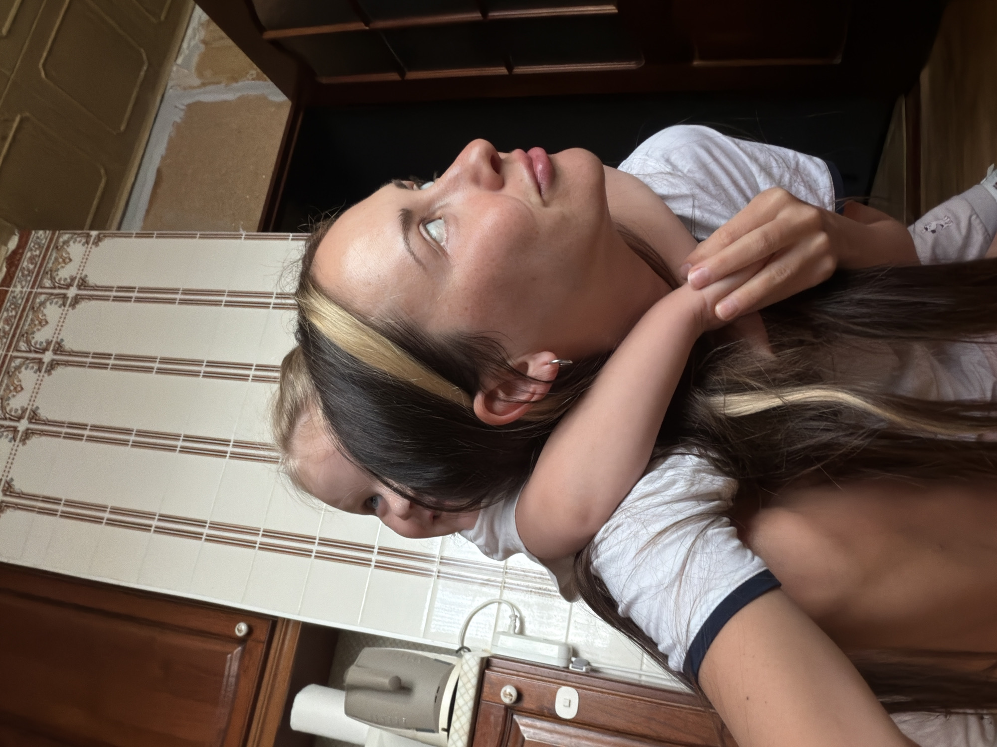 A woman is hugging a child. They are both wearing white t-shirts. The woman has long brown hair with blonde highlights. The child is nestled close to the woman's neck and back. The background appears to be a bathroom, with tiled walls and wooden cabinets. The location is not discernible as Khimki, Russia from the image.