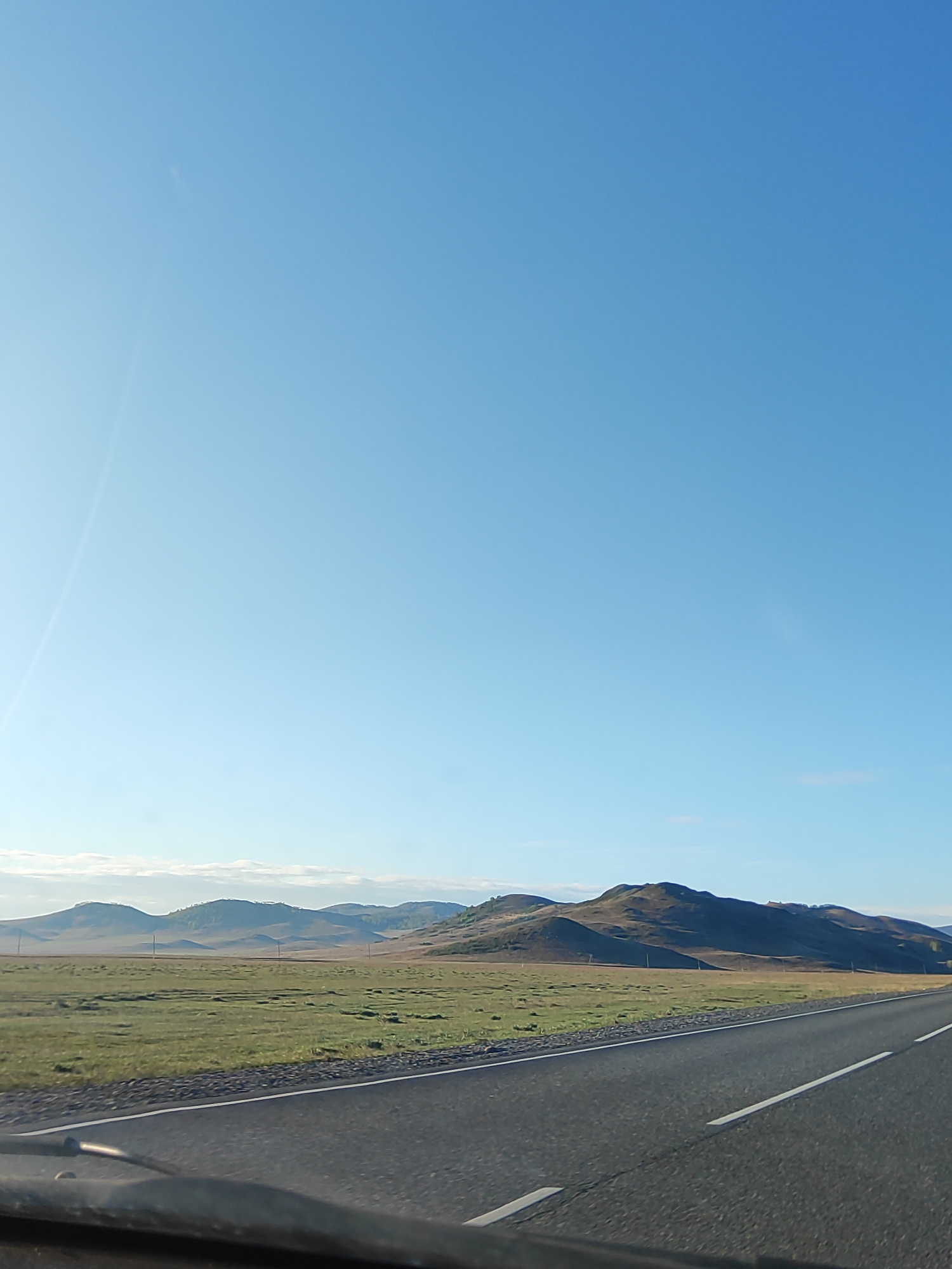 The image depicts a landscape with a road in the foreground. The road is asphalt and has white lines dividing the lanes. Beyond the road is a grassy field with rolling hills in the distance. The hills are brown and green with some trees. The sky is clear and blue. The scene appears to be taken from inside a vehicle, as part of the dashboard is visible. The location appears to be rural and sparsely populated.
