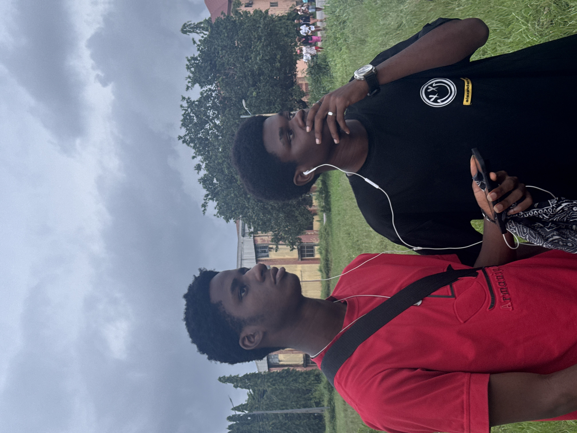 Two young Black men are standing in a grassy outdoor area, likely on the grounds of a university or a public park in Ihiagwa, Nigeria. The sky is overcast with thick, grey clouds, suggesting the possibility of rain.
The man on the left, closer to the camera, is wearing a red t-shirt with a subtle embossed logo that appears to be Armani. He has short, dark, curly hair and is wearing white earbuds. A black strap crosses his chest from his right shoulder to his left hip, likely belonging to a bag. His gaze is directed to his left.
The man on the right is wearing a black t-shirt with a white circular graphic of a smiley face with crossed-out eyes, and a yellow rectangular label with text that appears to read "alexander". He also has short, dark, curly hair and is wearing white earbuds. He is holding a smartphone in his left hand and has his right hand near his mouth, with a ring on his right ring finger and a watch on his right wrist. He is looking to his right.
In the background, there is a light-colored, multi-story building with visible windows, possibly an educational institution. There are also tall, slender trees lining the left side of the frame and a larger, dense tree behind the two men. Further in the background, several people, appearing to be walking, are visible. The ground is covered in green grass, which appears somewhat unkempt. A utility pole with a light fixture is visible on the far left.