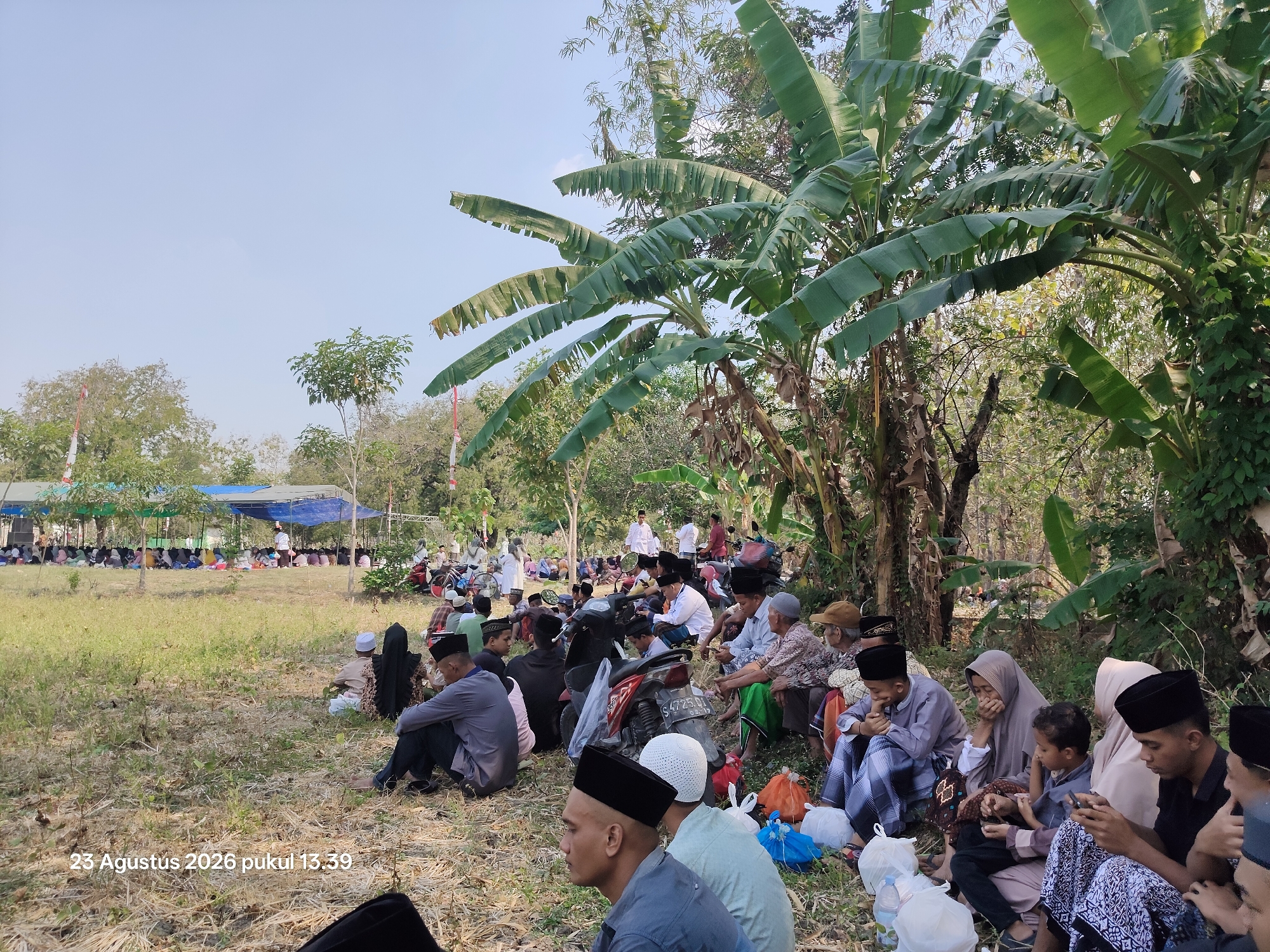 In Padang Mentoyo, Indonesia, on August 23, 2026, at 13:39, a group of people are gathered outdoors under a bright sky. The scene appears to be a community event, possibly religious, taking place in a grassy, open area with scattered trees and vegetation, including prominent banana trees.
Many individuals are seated on the ground, some in prayer or listening attentively. They are dressed in traditional Islamic attire, including peci hats and sarongs, with some women wearing hijabs. The crowd consists of men, women, and children.
In the background, temporary shelters with blue and green tarpaulins are set up, likely providing shade for the attendees. Several motorcycles are parked among the people, indicating transportation to the location. Bags and water bottles are visible on the ground, suggesting people are prepared for an extended gathering. The overall atmosphere suggests a communal and peaceful gathering.
