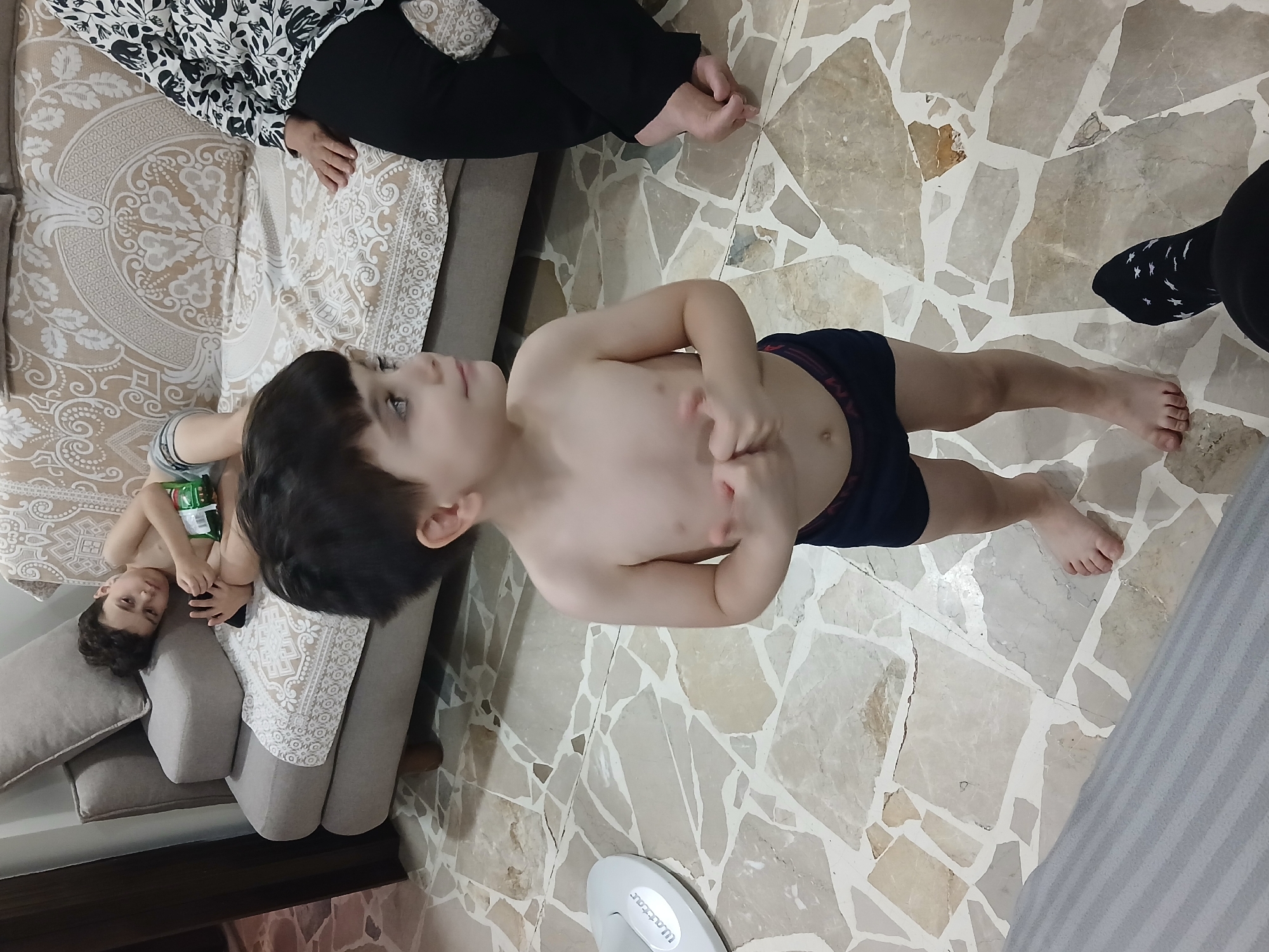 A young boy, wearing dark blue boxers, stands on a marble tiled floor, looking to his right. He has short, dark hair and is shirtless. His hands are clasped in front of his chest. In the background, an older boy lies on a couch, holding a bag of chips and looking at a phone. A woman, wearing a black and white patterned top and black pants, sits on the couch to the right. The environment appears to be an indoor living area. The image was taken in Barza Municipality, Syria.