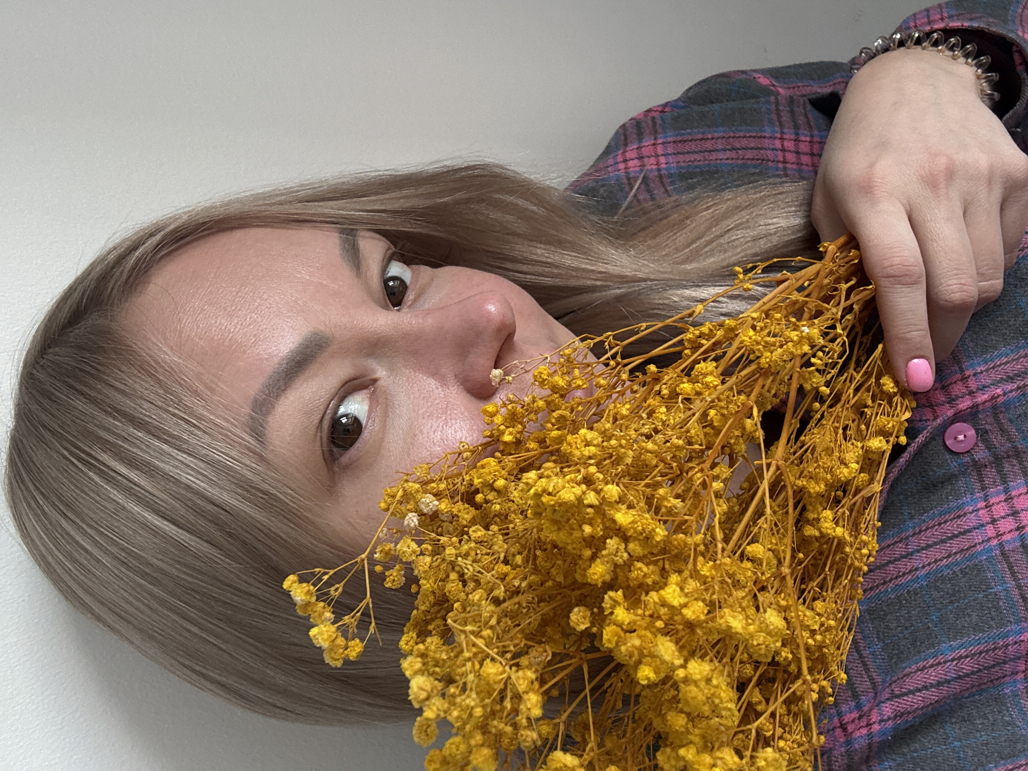 A woman with blonde hair and fair skin is holding a bouquet of dried yellow flowers. She is wearing a plaid shirt with pink and grey hues. Her right hand is holding the flowers, and she has a spiral hair tie on her wrist. Her right pinky finger is painted with pink nail polish. The background is a plain white wall.
