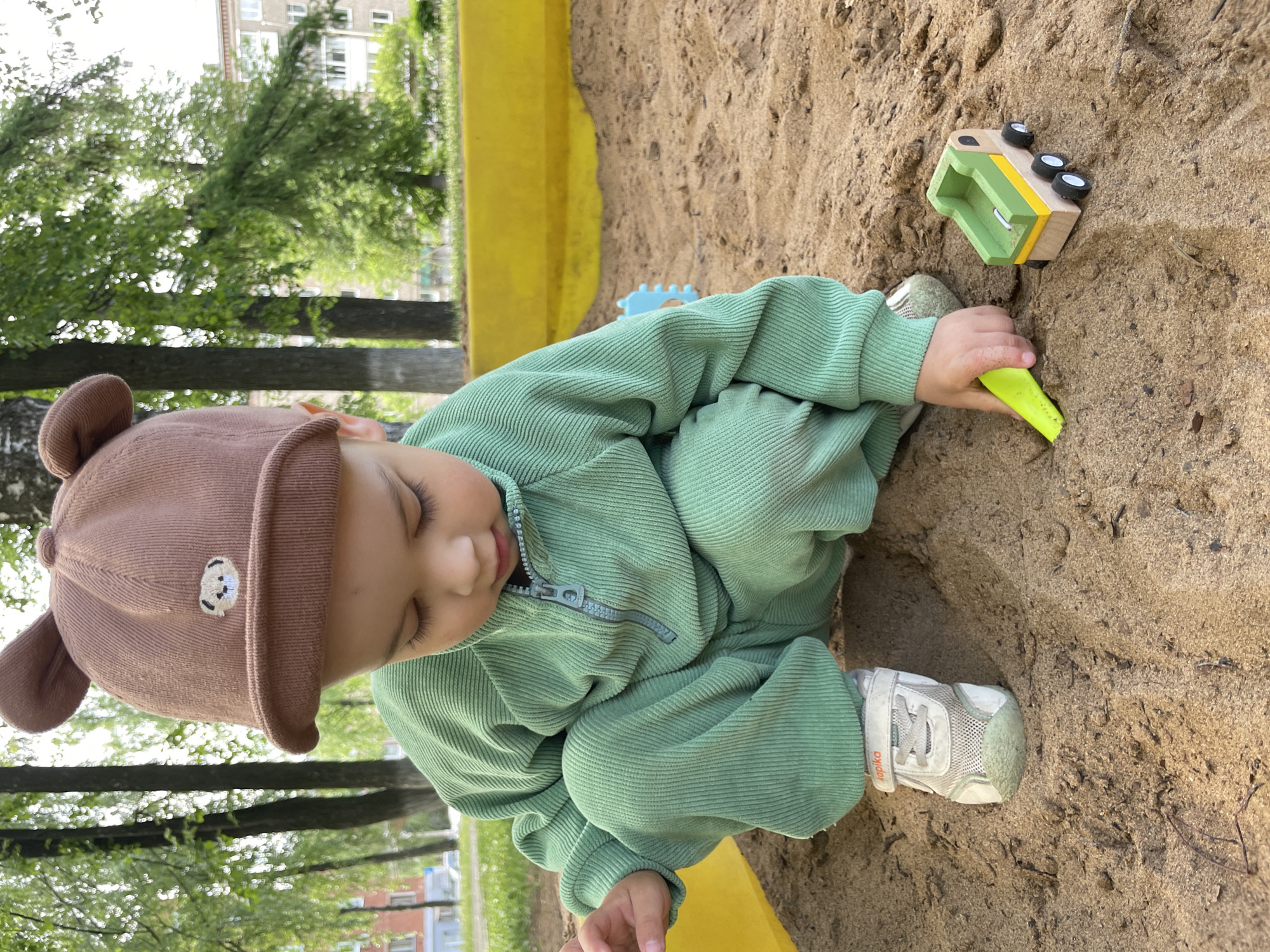 A young child wearing a brown hat with bear ears and a light green zip-up outfit is sitting in a sandbox. The child is crouched, holding a yellow plastic shovel and appears to be playing with a toy wooden truck. The sandbox has yellow edging. The background includes trees and a building. The city is Izhevsk, Russia.