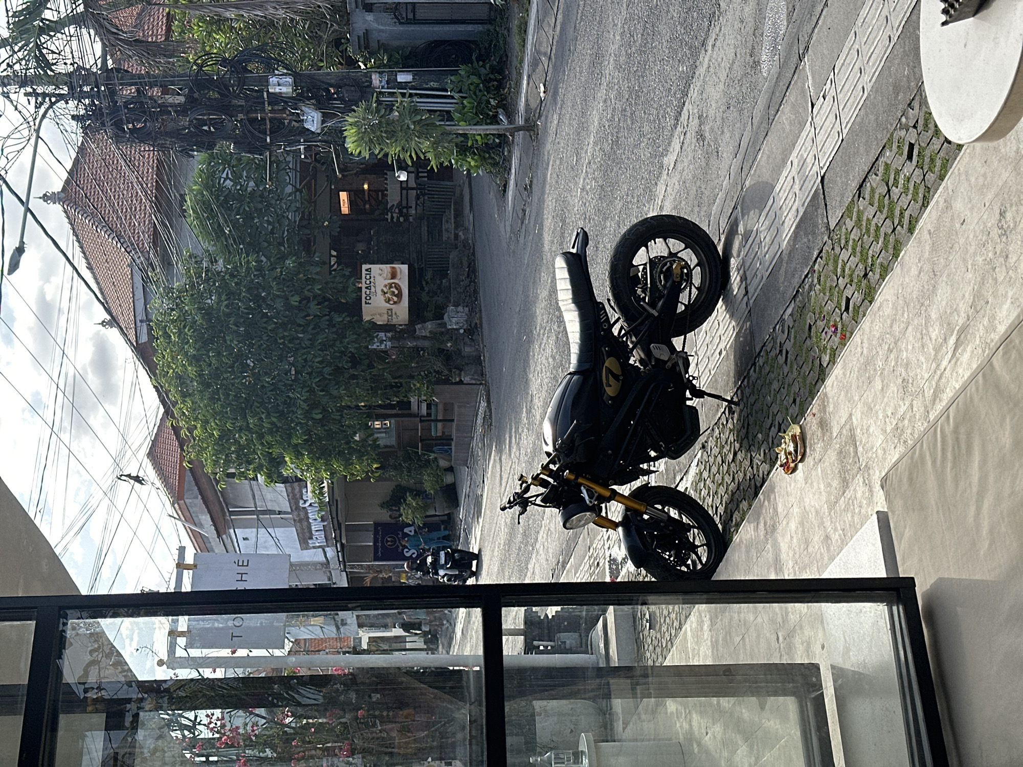 This is an outdoor, eye-level, medium shot of a street scene in Mengwi, Indonesia. In the foreground, a black motorcycle with gold accents is parked on a paved walkway. The motorcycle has a yellow number "7" on its side. To the left, a modern building with large glass windows and dark frames dominates the foreground. Through the windows, potted plants with pink flowers are visible. A white table with a light gray cushion is in the lower right corner.
The street is asphalt, and on the right side, there is a sidewalk with a tactile paving strip. The street is lined with buildings, including one with a red tile roof and intricate architectural details. A large tree with lush green foliage stands near the center of the frame. Electrical wires and a utility pole with a tangle of cables are prominent on the right side of the street.
In the mid-ground, people are visible on the street. A person on a motorcycle is riding down the street. Two individuals are standing on the sidewalk in front of a building with a sign that reads "FOCACCIA." Another sign above a storefront reads "Harmony Sang." A white sign with black text on a pole says "TO CHÉ."
The lighting suggests it is daytime, with sunlight casting shadows. The overall scene depicts a typical street in a tropical urban environment.