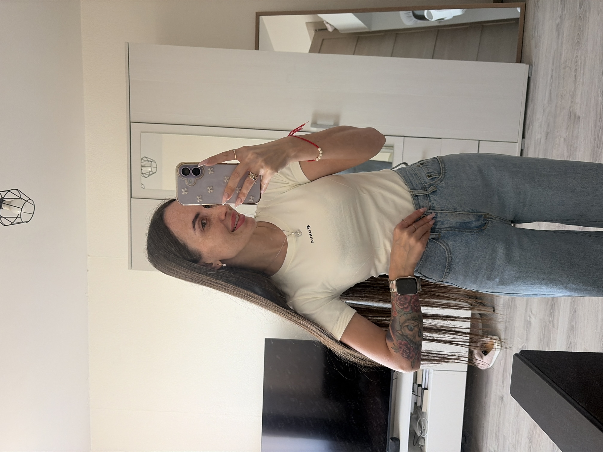 A woman is captured in a mirror selfie, holding an iPhone with a light purple case adorned with white floral patterns. She has long, dark brown hair falling over her shoulder and a pleasant, slight smile. She is dressed in a cream-colored, short-sleeved t-shirt with the word "СПЯЩАЯ" (Spyashchaya, meaning "sleeping" in Russian) printed on it, paired with light-wash blue jeans.
Her accessories include a silver Apple Watch on her left wrist, a red string bracelet with white beads on her right wrist, a silver necklace with a pendant, and multiple rings on her fingers. A detailed, colorful tattoo featuring an eye and roses covers her right forearm. Her fingernails are manicured with a light-colored polish.
The indoor setting appears to be a well-lit room in Saint Petersburg, Russia, likely during the daytime. Behind her, a large white wardrobe or cabinet is reflected in the mirror. The floor is made of light-colored wood. A dark TV screen is visible to her left, and a black metal light fixture hangs on the wall in the reflected background. The overall scene suggests a clean and modern interior.
