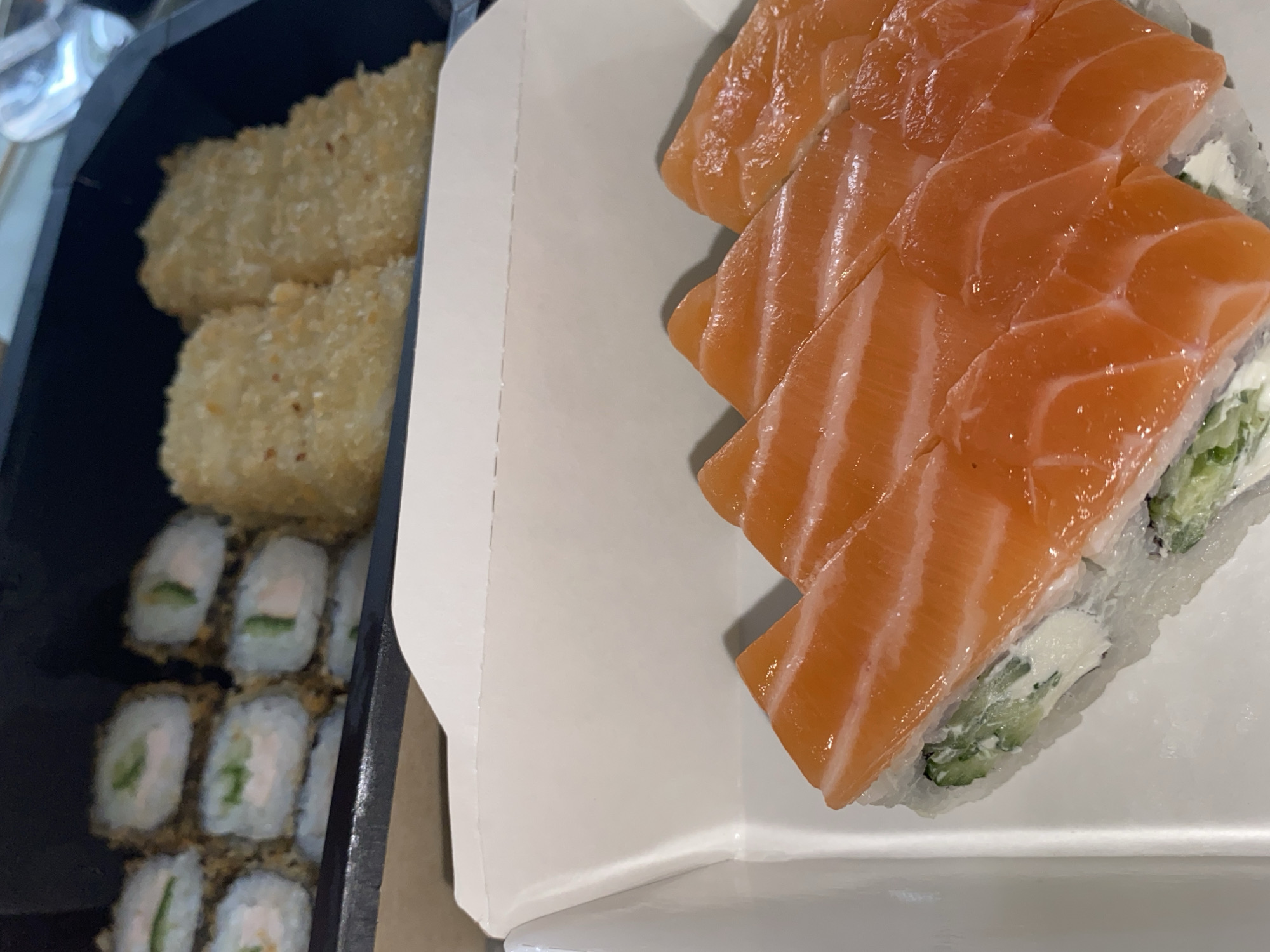 The image shows two containers of sushi. The first container, black, contains deep-fried sushi rolls and other golden-fried pieces. The second container, white, contains sushi rolls topped with salmon. The containers are positioned adjacent to each other on a flat surface. Location information is not present in the image.