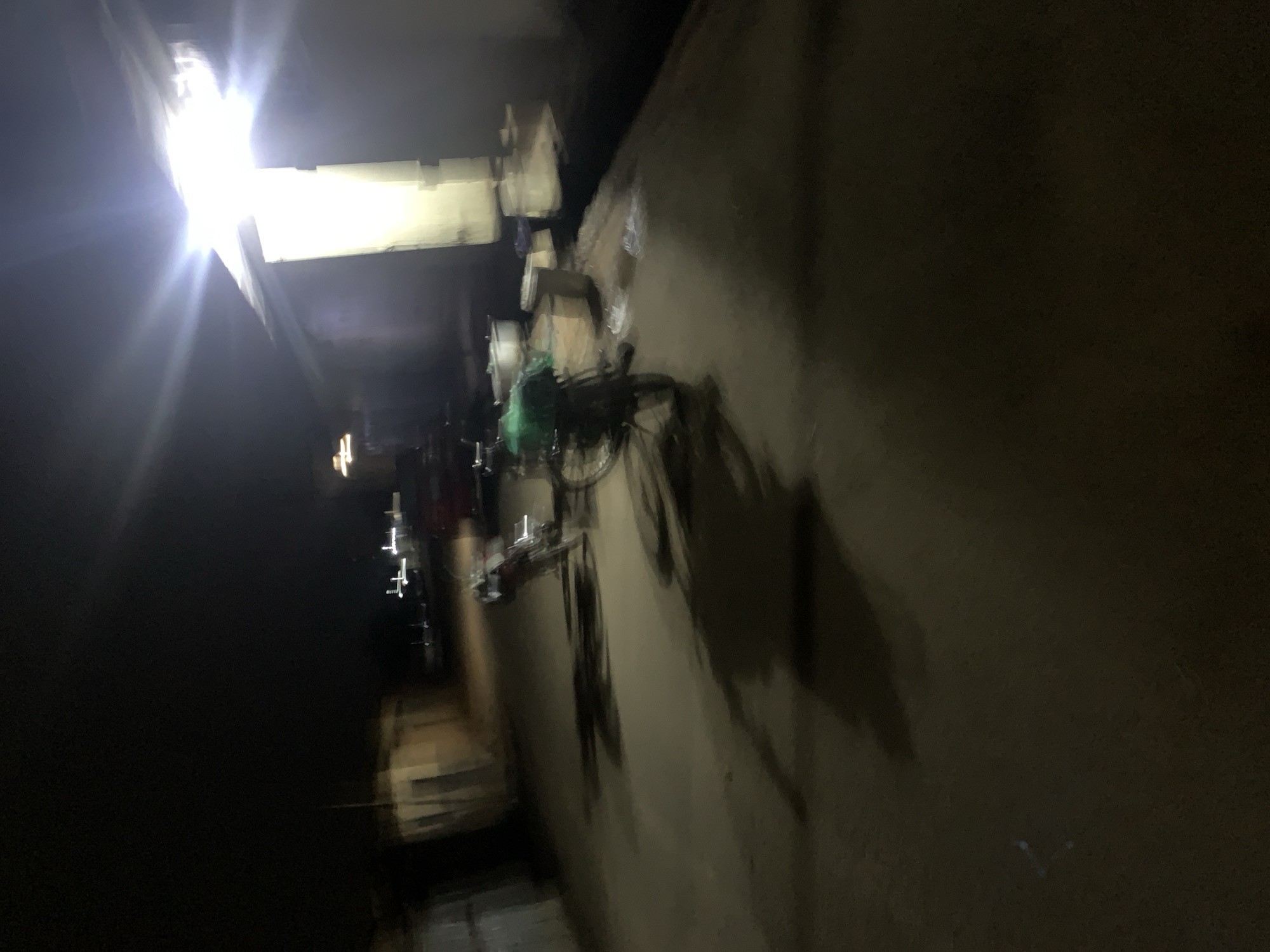 The image depicts a dimly lit outdoor scene, possibly at night, with a bright light source in the upper right quadrant creating lens flare and washing out details. In the foreground and mid-ground, there are several bicycles, some with green coverings, suggesting they are loaded or parked. The ground appears to be paved or a dirt path. In the background, there are structures that could be buildings or stalls. The overall impression is one of an urban or semi-urban environment at night. The image is blurry and lacking in detail, making precise identification of objects and the environment challenging. There are no clearly visible people. The context of Gura Top, Nigeria, is not evident from the visual information in the image itself.
