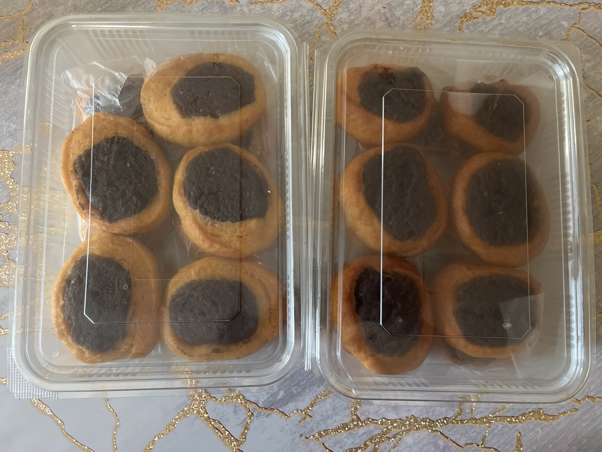 Two clear plastic containers of baked goods sit on a surface with a gold and grey marbled pattern. The top container holds six round pastries, each with a golden-brown crust and a dark brown filling in the center. The bottom container holds eight of the same pastries. The containers appear to be stacked vertically. The lighting suggests an indoor setting, likely a kitchen or dining area. No people are visible in the image.