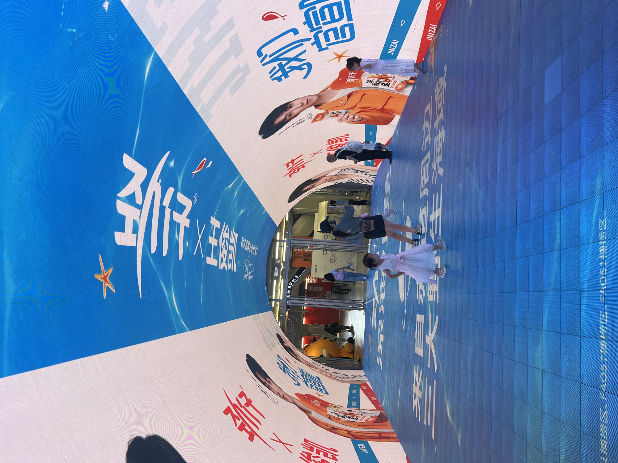 An outdoor pedestrian tunnel in Yuzhong District, China, is covered by a large, curved LED screen displaying a vibrant blue and white design with Chinese text and images. The screen features a starfish and a small fish graphic. On the left side of the tunnel, large red Chinese characters are displayed alongside images of a male celebrity wearing an orange jacket. On the right side, blue Chinese characters, also with a celebrity image and the text "JINZAI," are visible. The tunnel floor is also a large LED screen, displaying a blue grid pattern with white Chinese text that reads "来自XX的海域" (meaning "From the seas of XX"). Several people are present: a young girl in a white dress is walking towards the camera, another young girl in a white dress is standing and looking to the right, a woman in a black hat and grey shirt is standing and holding a phone, a man in a black shirt and white backpack is walking away from the camera, and other individuals are visible in the background near an entrance. The overall scene suggests a promotional event or a digitally enhanced public space.