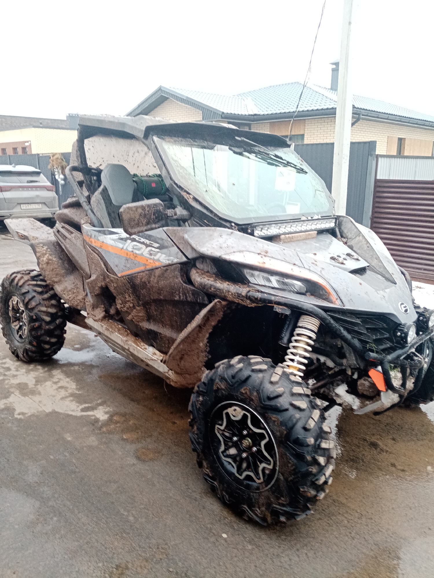 The image depicts a dirty gray and orange CFMOTO ZFORCE 1000 side-by-side ATV parked on a wet concrete surface in front of a residential area in Stary Chultem, Russia. The ATV is heavily covered in mud, particularly on the tires and body panels. A brick house with a gray roof is visible in the background, along with a silver SUV parked behind a gray fence.