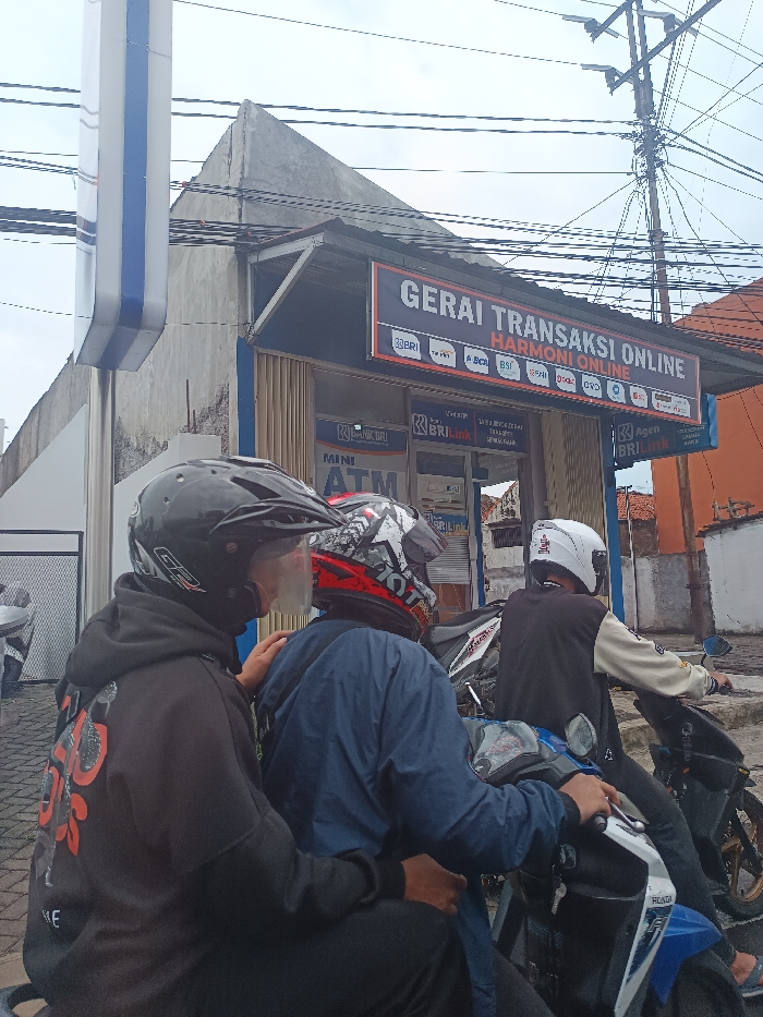 The image depicts an urban street scene in Cikaroya, Indonesia, under an overcast sky. In the foreground, three individuals on two motorcycles are positioned, facing away from the viewer. On the left, two individuals occupy a single motorcycle: the rear passenger, wearing a black full-face helmet with a clear visor and a black hoodie, places their left hand on the right shoulder of the driver. The driver wears a red, black, and white helmet and a dark blue jacket. To their right, a third individual sits alone on a blue and white scooter-style motorcycle, wearing a white helmet and a black and cream/white jacket, with hands on the handlebars.
In the background, a building houses a business named "GERAI TRANSAKSI ONLINE HARMONI ONLINE," indicated by a large sign displaying logos of various Indonesian banks including BRI, Mandiri, BNI, BCA, BSI, OCBC, Permata, Danamon, BTN, Agrodana, and Panin. The storefront below this sign features glass doors and windows, with "BANK BRI," "BRI Link," and "MINI ATM" signage. To the immediate left of the building, a tall, vertical blue and white pole structure is partially visible. Numerous tangled utility cables and power lines crisscross overhead against the sky. An orange-colored building is partially visible to the far right of the scene. The ground in the foreground appears to be paved.