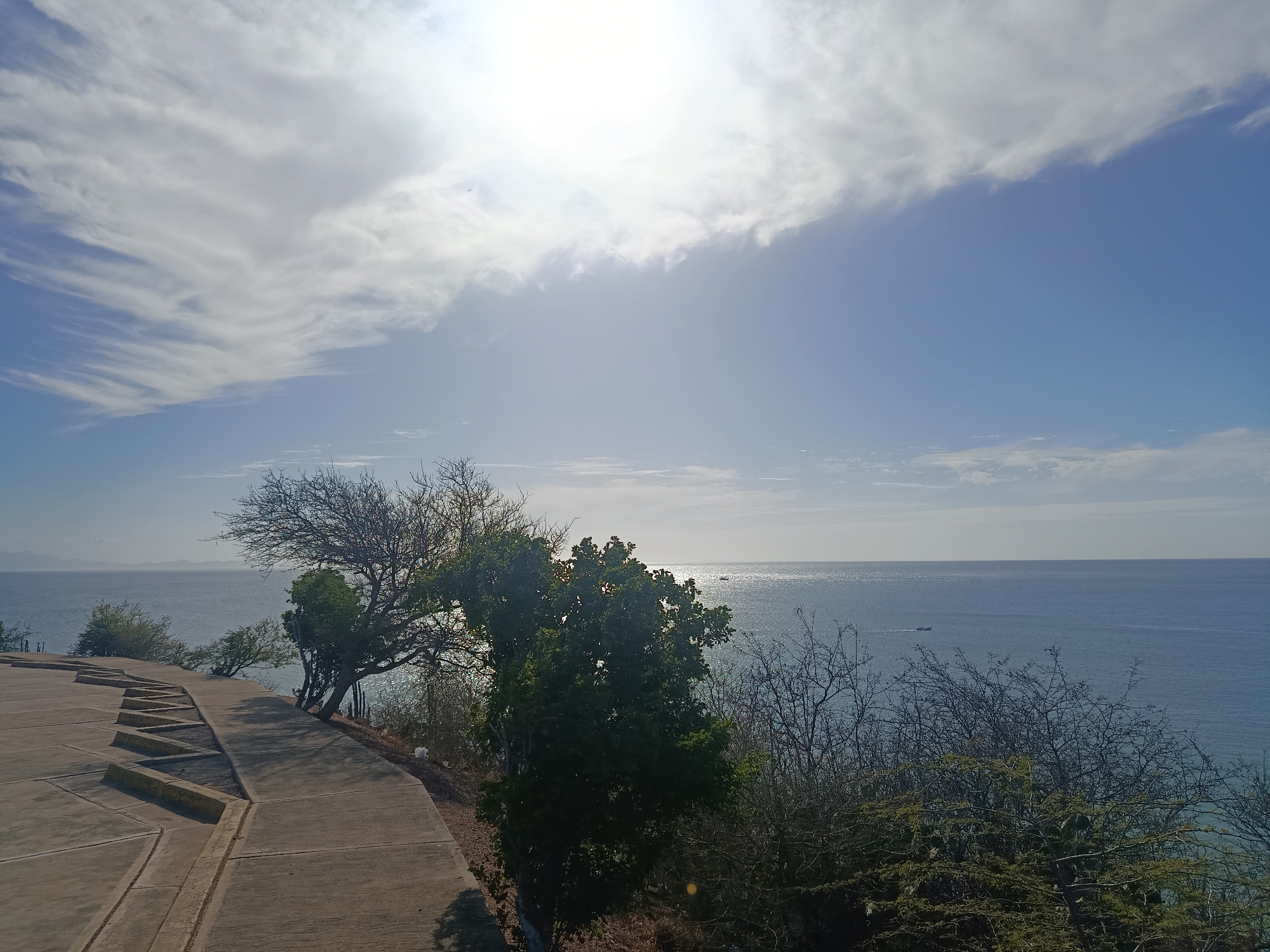 The image depicts a coastal scene in Juan Griego, Venezuela. A concrete walkway with stepped levels runs along the edge of a cliff. Beyond the walkway, there are trees and bushes growing on the cliffside. The view overlooks the ocean, which stretches to the horizon under a blue sky with scattered clouds. The sun is high in the sky, creating a bright, slightly overexposed effect in the clouds. A few distant boats are visible on the water.