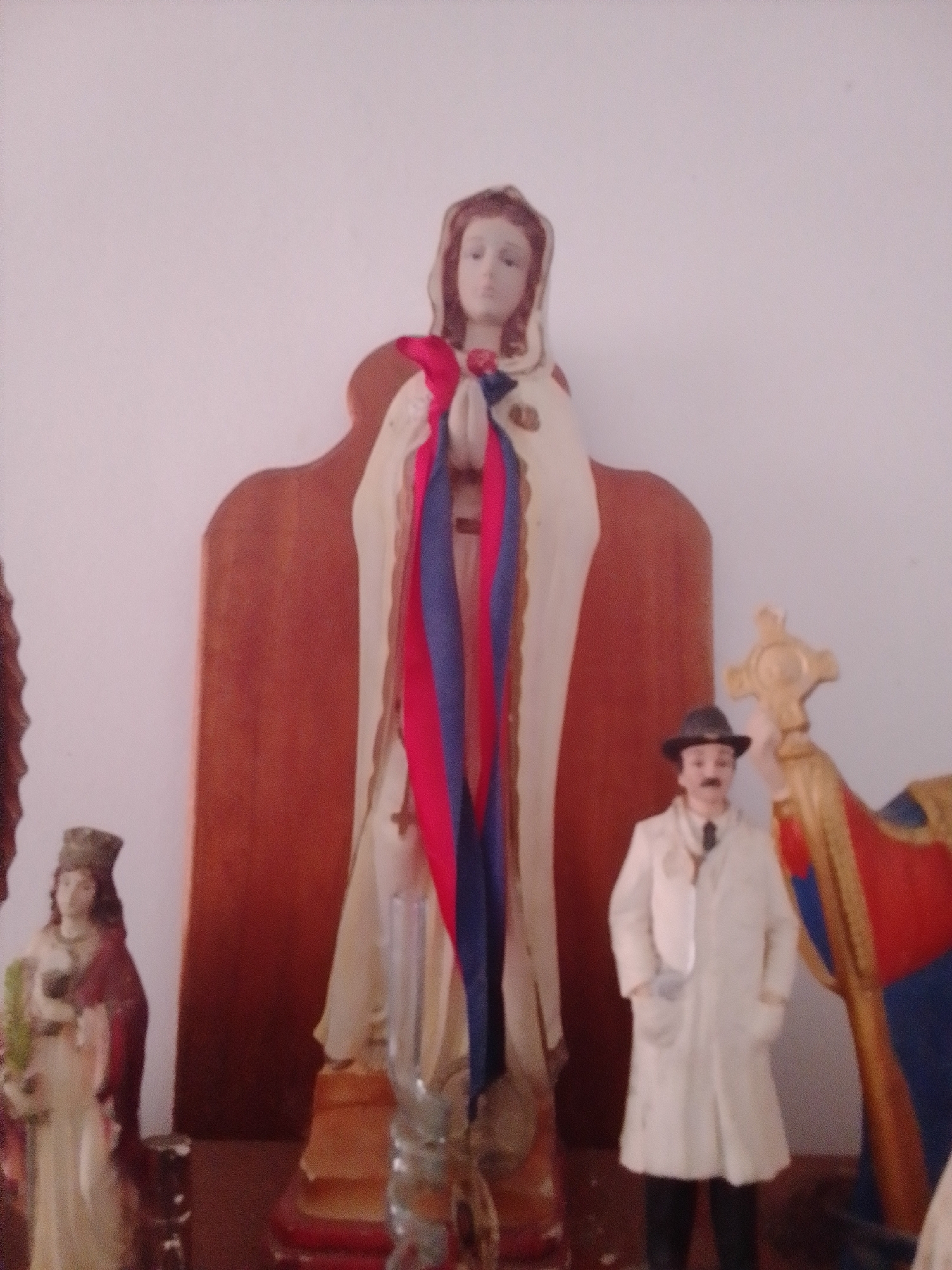 The image displays a collection of religious figurines displayed against a plain wall. The central figure is a tall statue of a woman, likely the Virgin Mary, wearing a white robe adorned with red and blue sashes. To her right stands a figurine of a man in a white coat, black hat, and tie, with his hands in his pockets. To the far left, another figurine of a woman in a red and beige dress is visible. Behind the central figurines, a wooden stand with a curved top is present. To the right of the man figurine, a golden cross with red and blue fabric is partially visible. The scene is lit by ambient light, casting soft shadows. The location is identified as La Tagua, Venezuela.