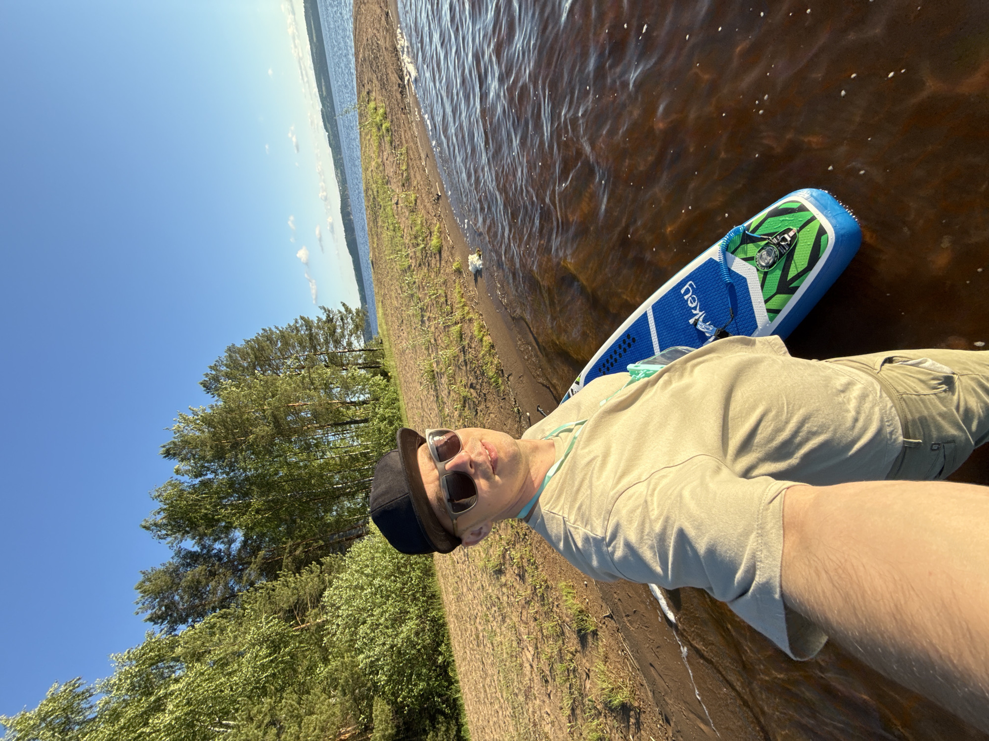 A man wearing a beige t-shirt, khaki pants, sunglasses, and a black baseball cap takes a selfie with a blue and green inflatable paddleboard at the edge of a body of water. The water is a dark reddish-brown color and the shore is sandy with some sparse green vegetation. Behind the shore, a dense forest with tall trees and green bushes is visible under a clear blue sky with a few scattered white clouds. The sun is shining, casting a warm light on the scene. The paddleboard has "Monkey" written on it in white letters. The image appears to be taken in the vicinity of Gora (Khokhlovskoe S/P), Russia, given the characteristic landscape of forests and lakes common to the region.