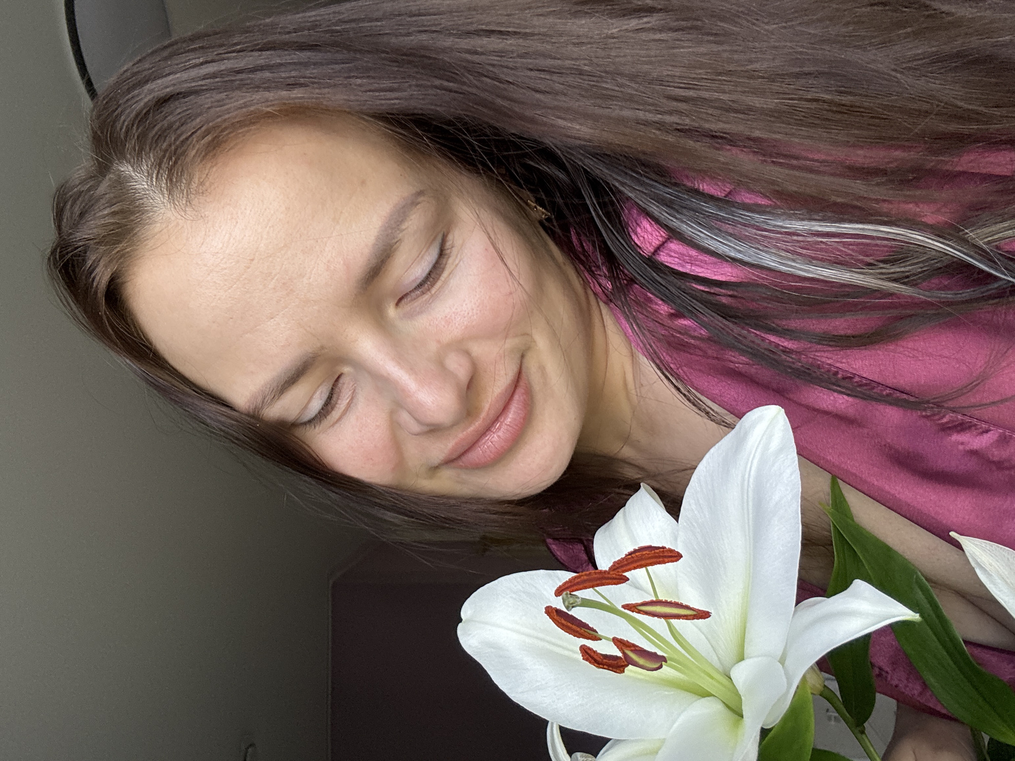 A woman with long, dark brown hair and closed eyes is shown in a close-up portrait. She is smiling softly and appears to be resting her head near a large white lily with prominent brown stamens. She is wearing a pink silk or satin top. The background is indistinct, but appears to be a wall. There is no evidence to place the location in Rusalevka, Russia.