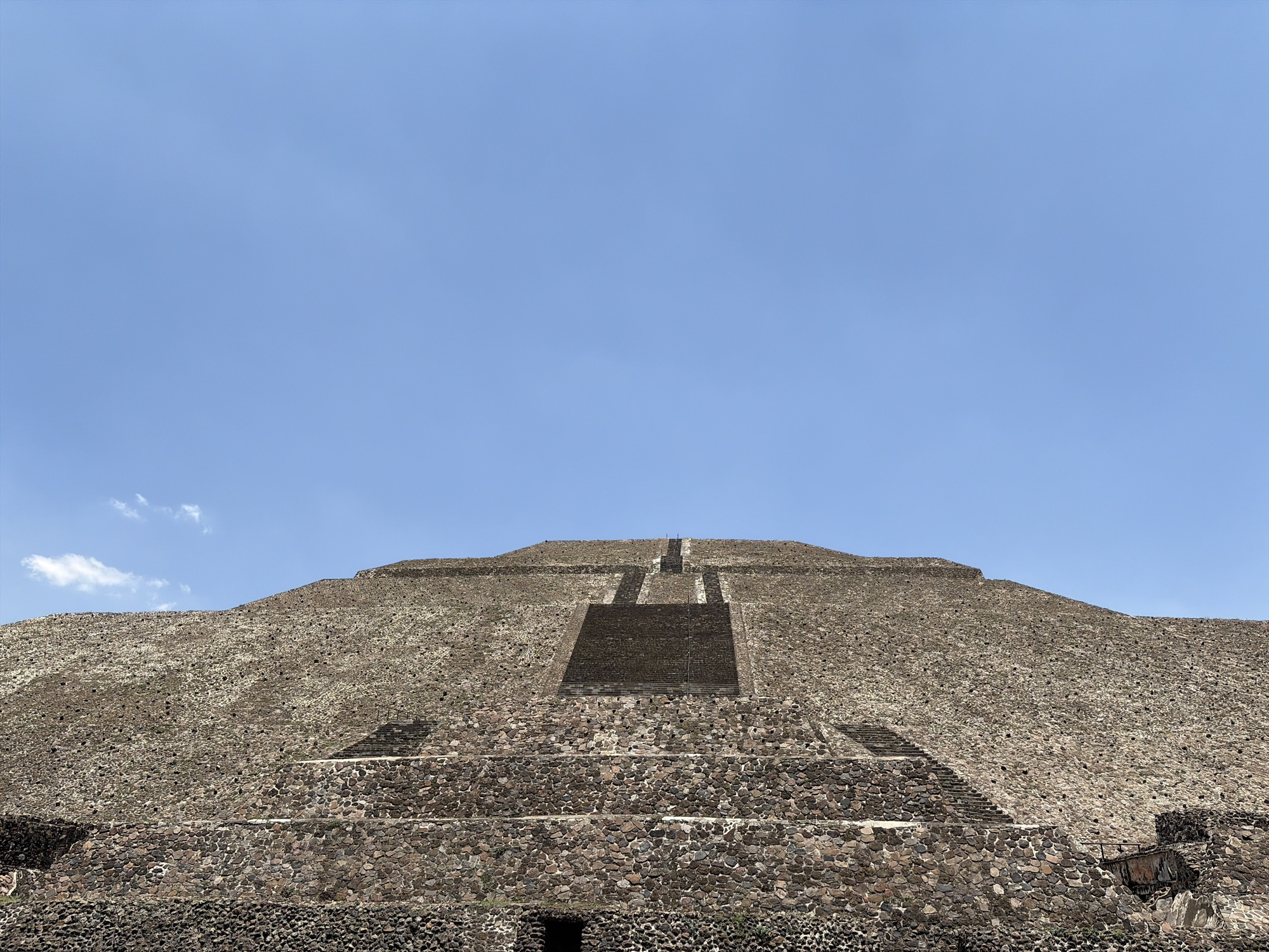 The image shows the Pyramid of the Sun in Teotihuacán, Mexico. The pyramid is constructed from dark-colored stones and features a central staircase leading to the summit. The sky above is a clear, light blue with a few wispy clouds.