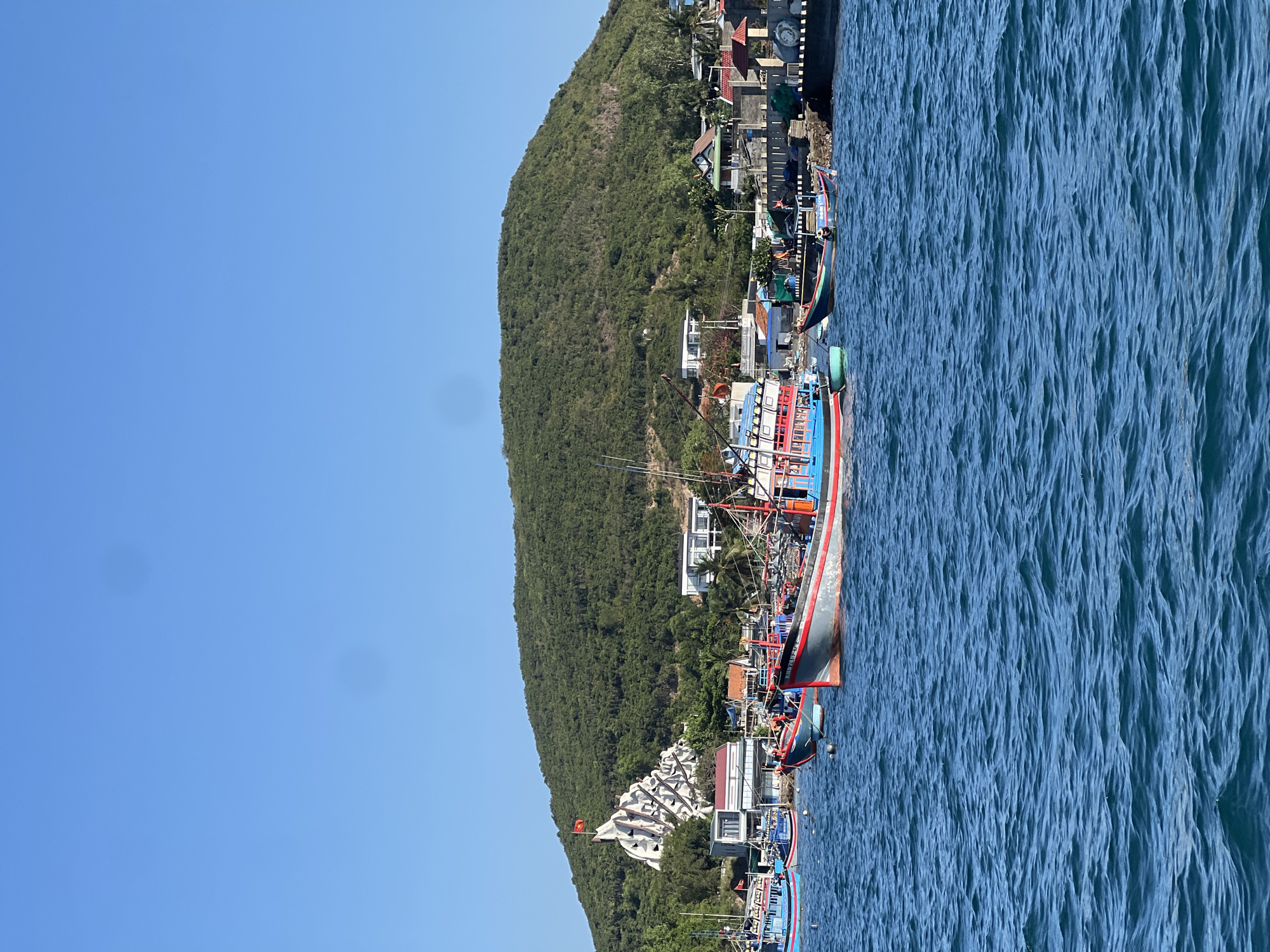 A vibrant, daytime scene depicts a coastline in Nha Trang Ward, Vietnam, with docked fishing boats and the adjacent terrain.
The foreground is dominated by the blue, slightly choppy water. Moored along the shore are several colorful fishing boats, notable for their size and traditional design. The boats display a mix of red, blue, and white paint schemes.
The terrain rises immediately behind the boats, densely covered with lush, green vegetation. The vegetation seems to be primarily trees and other dense foliage. Nestled amongst the greenery are a few buildings and structures, appearing to be residential or commercial, blending into the hillside.
Above the coastline, the sky is clear and bright blue. The image is composed with a strong emphasis on showcasing the boats, coastline, and the vibrant mix of human activity and natural environment.