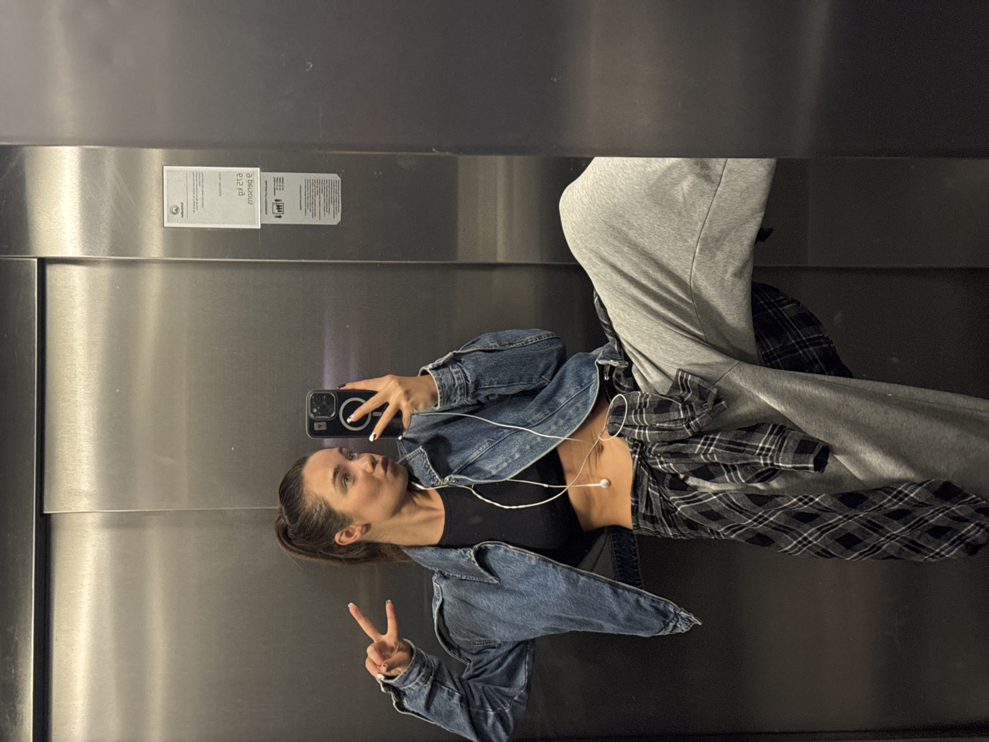 A young woman is captured in a mirror selfie inside a metallic elevator. She is positioned facing her reflection, with her head slightly tilted and a relaxed expression, making a peace sign with her left hand. Her right hand holds a dark-colored smartphone, possibly an iPhone, which is the device capturing the image. She is wearing a dark blue denim jacket over a black crop top, revealing her midriff. A black and white plaid shirt is tied around her waist, and she wears light grey sweatpants. White wired earbuds are visible, plugged into her phone and hanging down.
The elevator walls are made of brushed or polished stainless steel, reflecting the surroundings. On the upper portion of the wall to the left, an informational sticker or plaque is visible. Due to the reflection and angle, the text appears reversed and partially upside down. Identifiable elements include "e12 kg" (likely relating to weight or capacity, possibly "21 kg" if viewed correctly) and a logo that resembles "KONE," a well-known elevator company. Further small, illegible text likely provides instructions or technical details. The setting is an indoor, utilitarian space, typical of an elevator in an urban environment like Budva, Montenegro, as provided by the context. The woman's pose and gesture suggest a casual and confident mood.