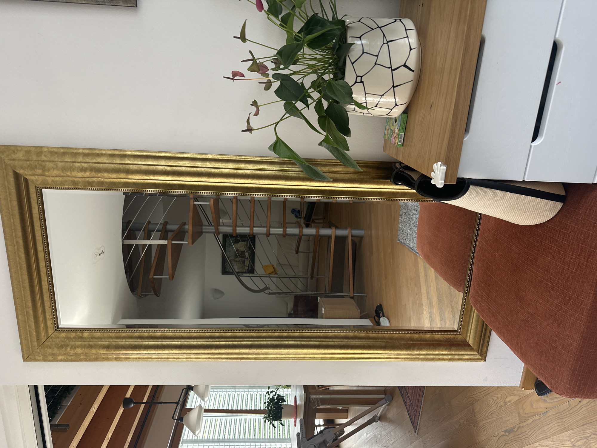 This image captures an interior scene, likely a residential hallway or living area. A tall, ornate gold-framed mirror dominates the left side of the frame, reflecting a modern spiral staircase with wooden steps and metal railings, as well as a framed picture on the wall. To the right of the mirror, a white chest of drawers with a wooden top is visible. On top of the chest sits a potted plant with dark green leaves and purplish-red flowers in a decorative white and black patterned pot. A light-colored woven bag with black trim leans against the side of the chest, and a small white object resembling a hand is attached to the edge of the chest. In the foreground, the corner of a rust-colored upholstered ottoman or bench is visible, positioned in front of the mirror. The floor is made of light-colored wood planks. The scene appears to be well-lit with natural light filtering in from unseen windows. There are no visible people in the image. The city of Trieste, Italy, is not depicted or implied in this image.