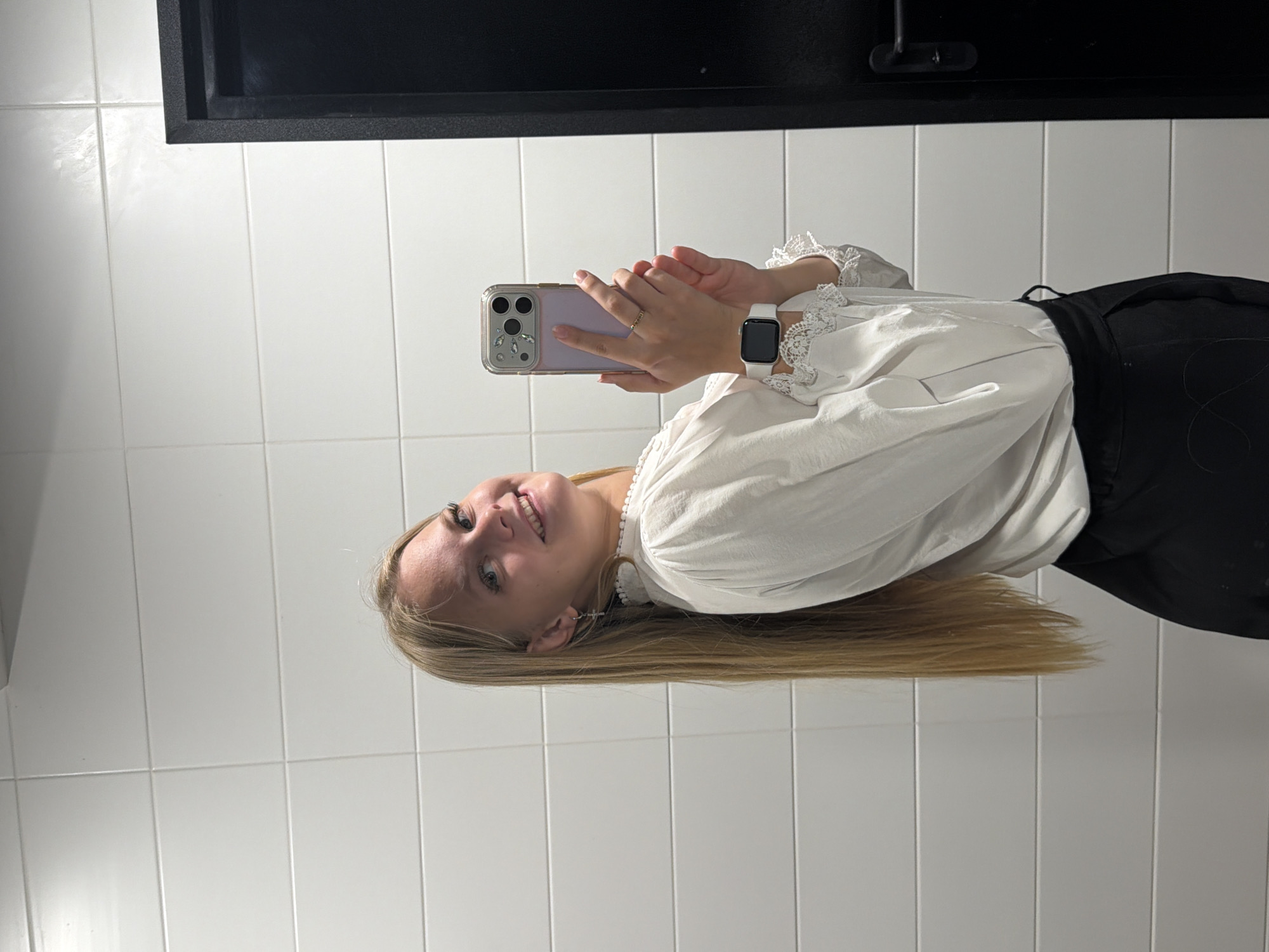 A young woman with long blonde hair takes a selfie in a restroom. She is wearing a white blouse with lace trim and black pants. She is holding a smartphone in her right hand, and has a smartwatch on her left wrist. The walls of the restroom are tiled with white square tiles. A black door is visible on the right side of the frame. The image is a horizontal shot, taken from a medium distance. The lighting is bright and even.