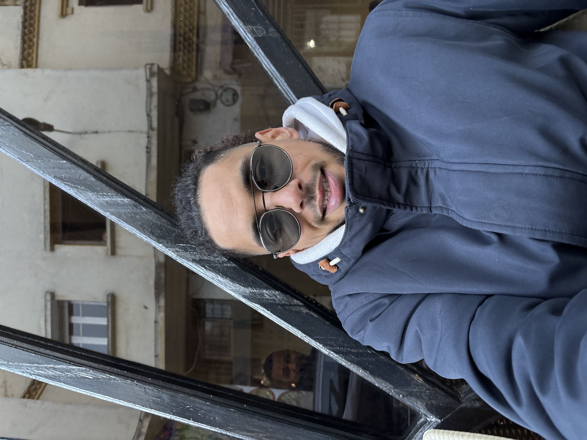 A man with curly dark hair and a mustache is seen smiling, wearing sunglasses and a blue jacket with a white hood. He appears to be leaning against a dark metal railing. Reflected in the glass behind the railing is a building with windows and architectural details, suggesting an urban environment. Another person is also reflected in the window. The setting could be a balcony or a window overlooking a city street. The location is identified as Tanger طنجة, Morocco.