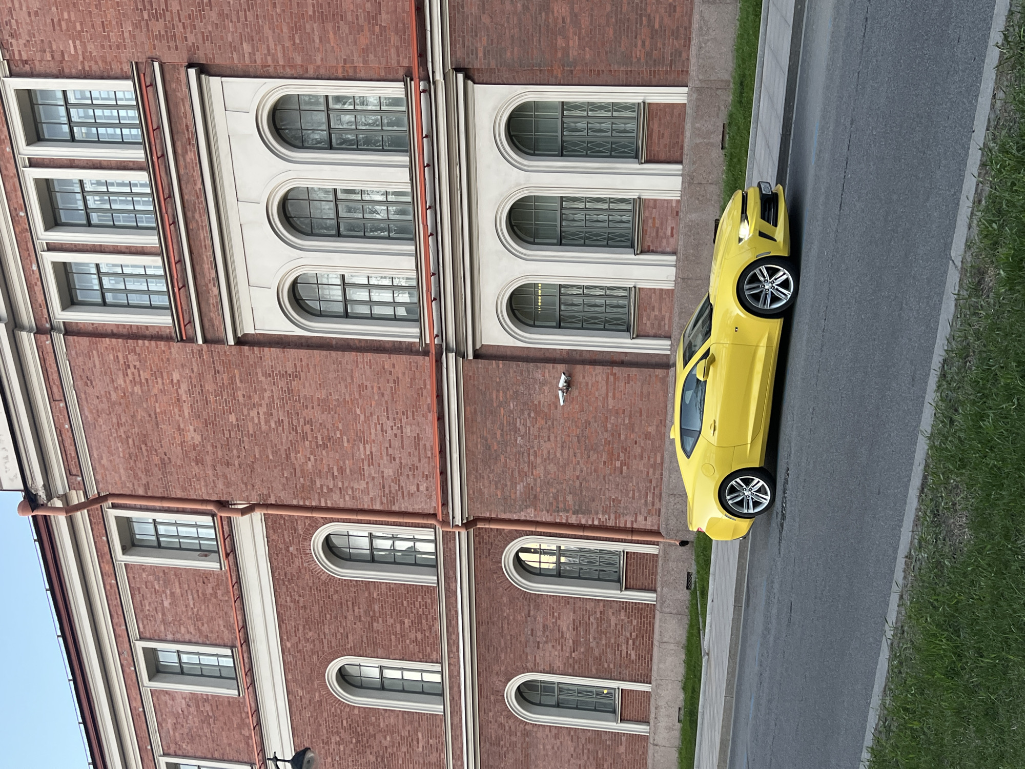 A yellow car is parked on the street in Saint Petersburg, Russia, next to a brick building. The building has multiple windows, some rectangular and some arched. There is grass growing along the edge of the sidewalk next to the street.