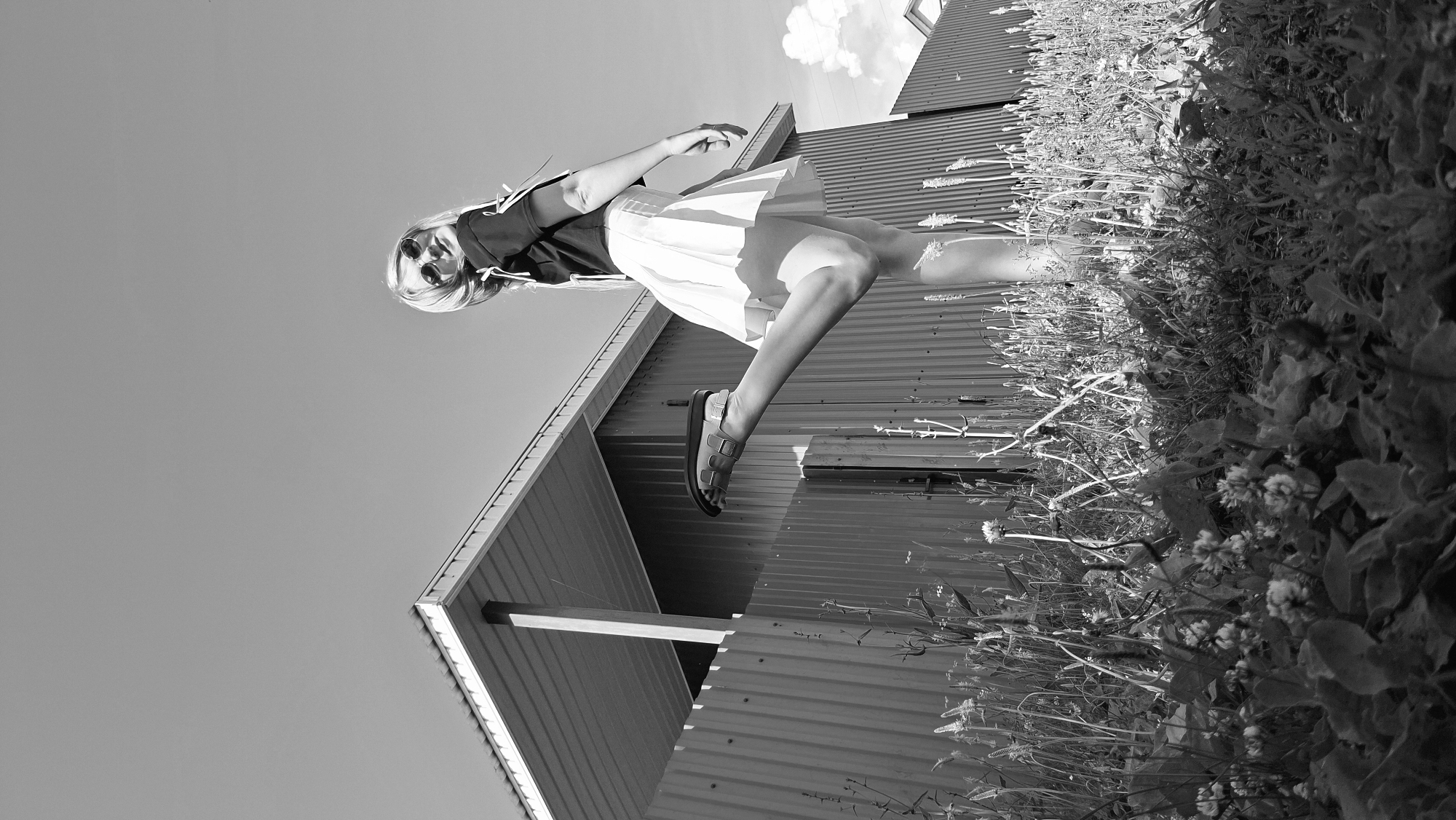 In a black and white photograph, a young woman stands with one leg raised, her foot wearing a sandal, in front of a corrugated metal fence and a building with a peaked roof. The woman has blonde hair and is wearing sunglasses, a dark top with decorative ties, and a light-colored pleated skirt. She is looking towards the camera with a smile. The scene appears to be outdoors, with grass and wildflowers in the foreground, and a clear sky with a few clouds in the background. The perspective is low-angle, looking up at the woman. The location is identified as Bolschoi Bukor, Russia.