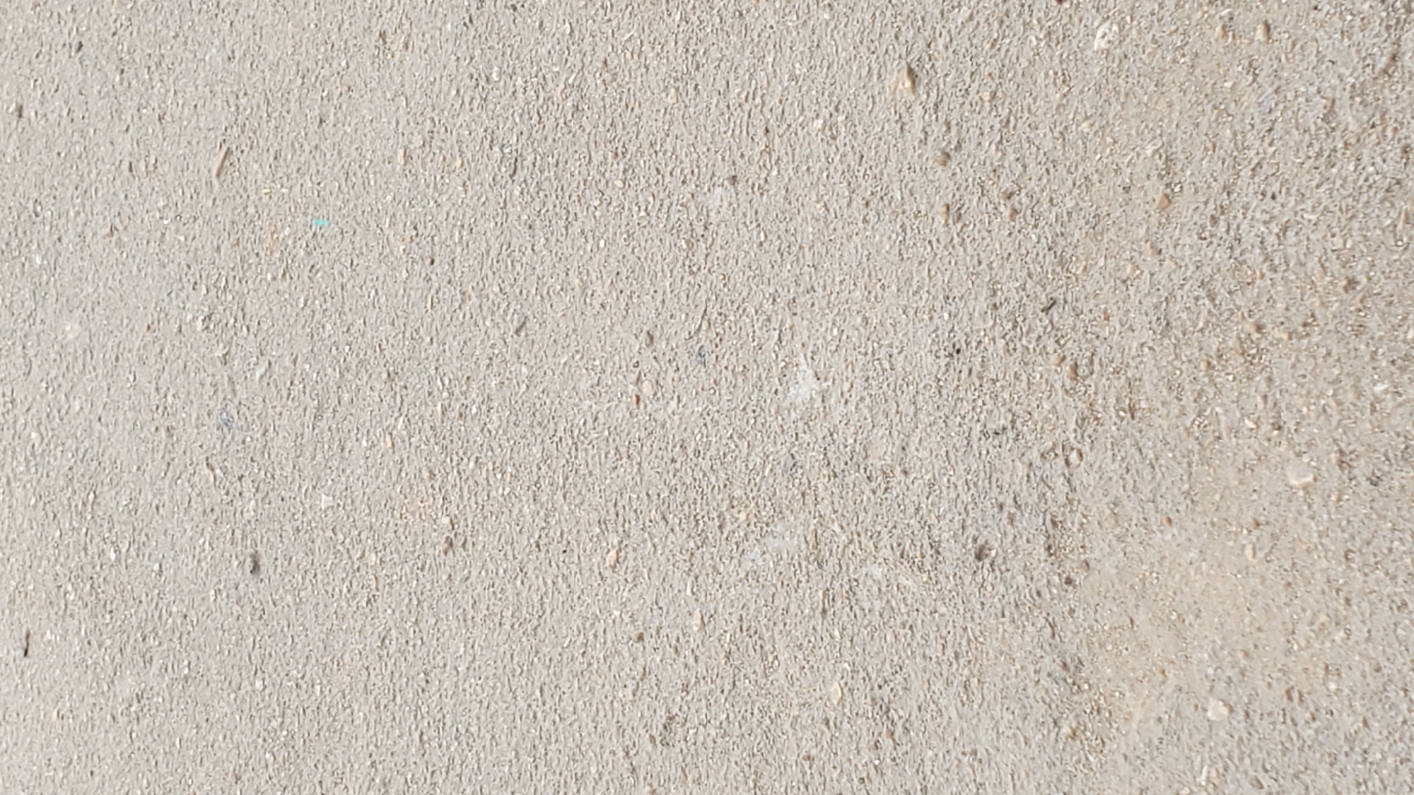 The image displays a close-up, top-down view of a light-colored, granular surface, likely asphalt or compacted gravel. The texture is characterized by a uniform distribution of small pebbles and sand particles. There are no discernible objects, people, or activities captured within the frame. The lighting suggests an overcast or diffused daylight condition. This scene could be a section of a road, pathway, or open ground in Jalingo, Nigeria.