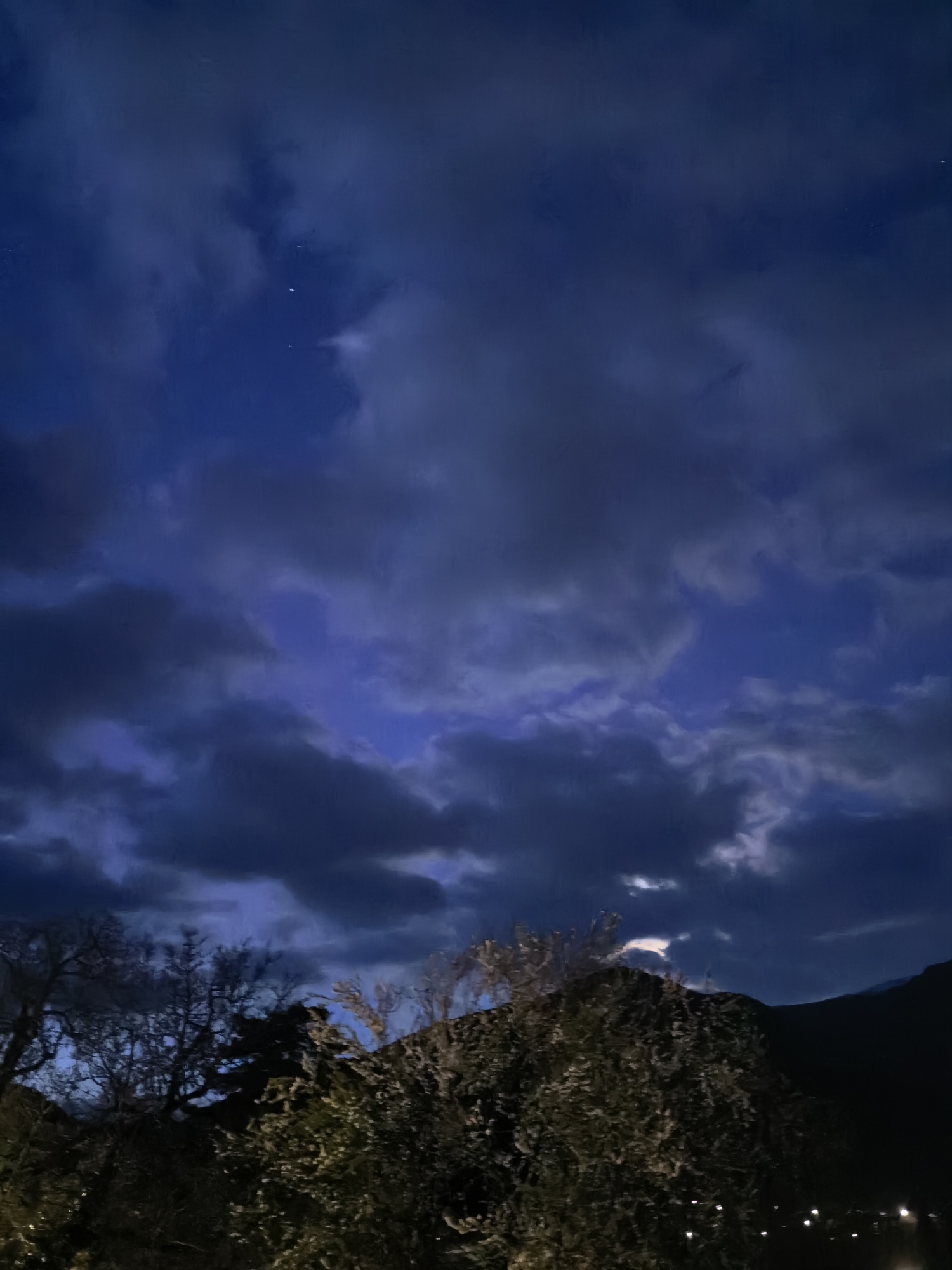 The image depicts a night scene in an unknown city. The sky is filled with dark, heavy clouds, with patches of a deep blue color peeking through. There are a few visible stars. In the foreground, there's a silhouette of a tree and a bush or hill covered in vegetation. In the distance, a dark hill or mountain can be seen, along with what appear to be street lights from the city.