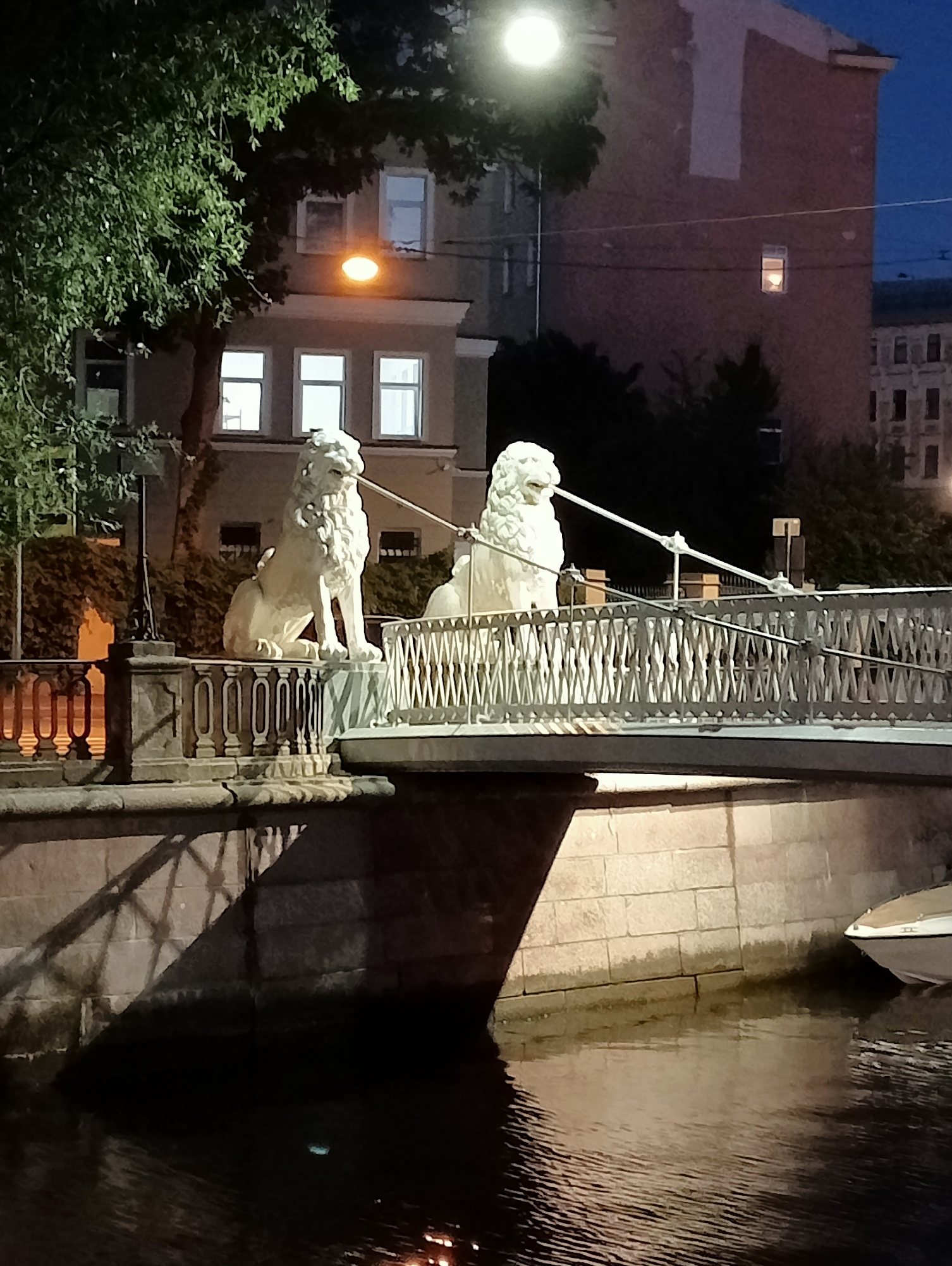 The image depicts the Lions Bridge (L'viny Most) in Saint Petersburg, Russia, at dusk or night. Two white lion statues are prominently featured, seated on either side of the bridge's entrance. The bridge itself has an ornate white metal railing with a crisscross pattern. Behind the bridge, a multi-story building with illuminated windows is visible. The bridge spans a dark body of water, likely a canal, with reflections of the bridge and lights on its surface. A portion of a white boat is visible on the right side, partially submerged in the water. Trees with dense foliage are present on the left side of the image, partially obscuring a building. The overall atmosphere is dimly lit, suggesting a nighttime scene in an urban environment.