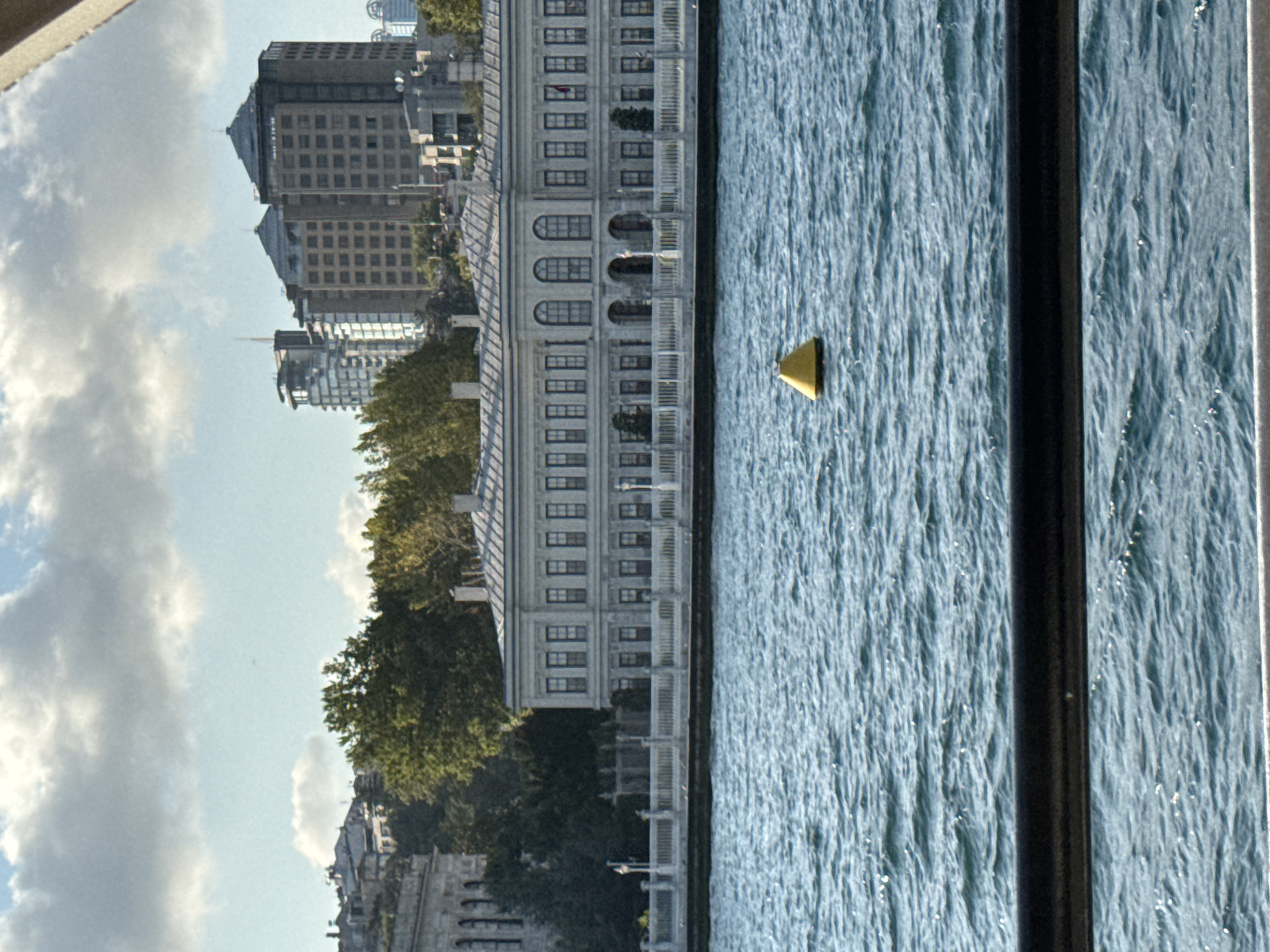 A wide shot captures the Bosphorus strait in Istanbul, Turkey, with a view of the Dolmabahçe Palace in Beşiktaş. The palace, a grand neoclassical structure with a long facade and numerous windows, stands prominently on the shore, bordered by a white ornate railing. Behind the palace, lush green trees and a cluster of modern buildings, including skyscrapers, rise against a sky filled with scattered clouds. In the foreground, the rippling blue water of the strait is dotted with sunlight reflections, and a bright yellow buoy floats in the middle distance. A dark metal railing runs across the very bottom of the frame, partially obscuring the view of the water. There are no people visible in the scene.