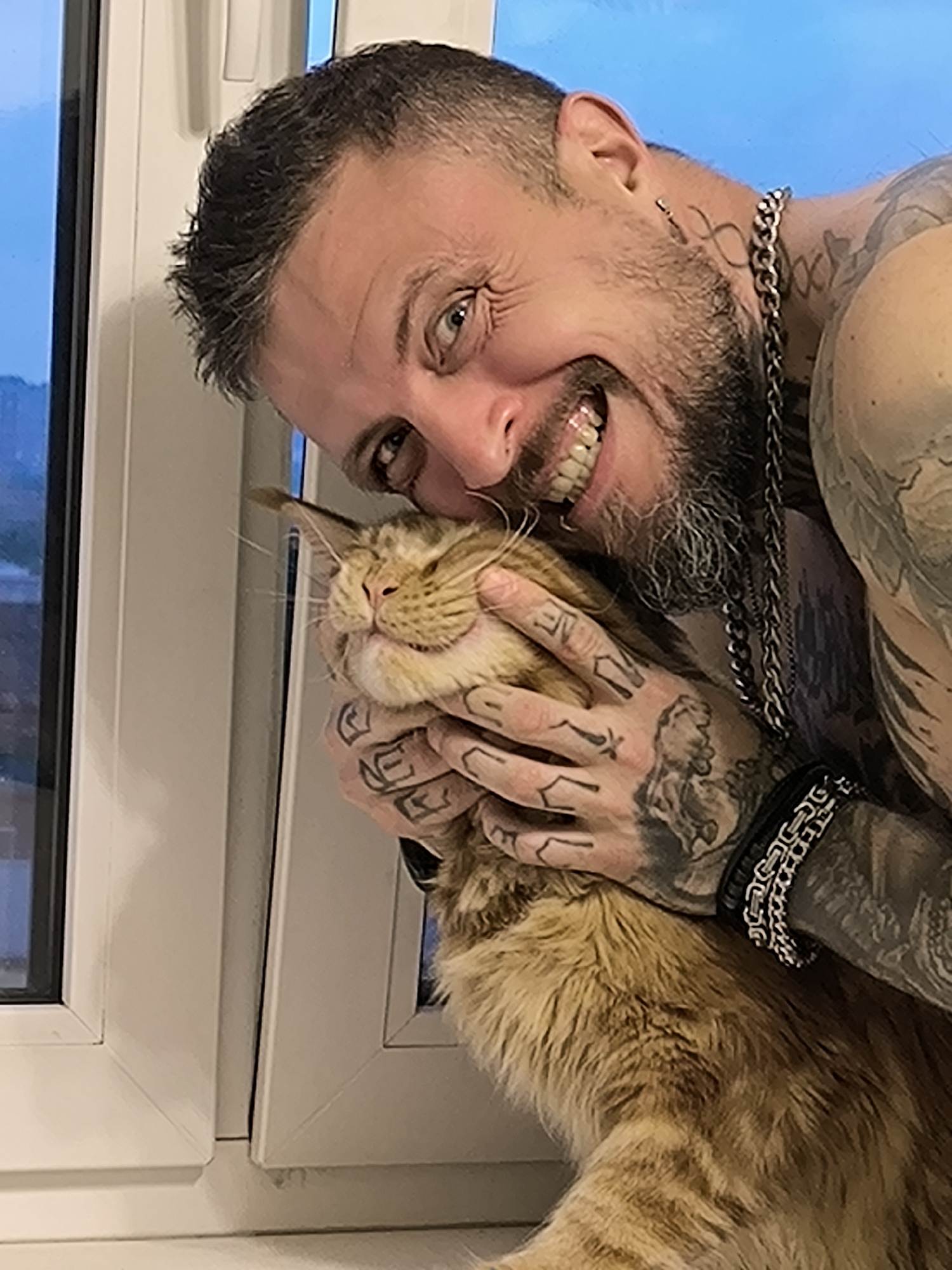 A shirtless, tattooed man with dark hair and a beard smiles widely, holding a fluffy orange cat. The man is leaning against a white window frame, and the cat is nuzzling into his face. The background appears to be an interior space with a window showing a blurred, dusky sky. The image is likely a personal photograph, possibly taken in a residential setting in Saint Petersburg, Russia, given the style of the window frame.