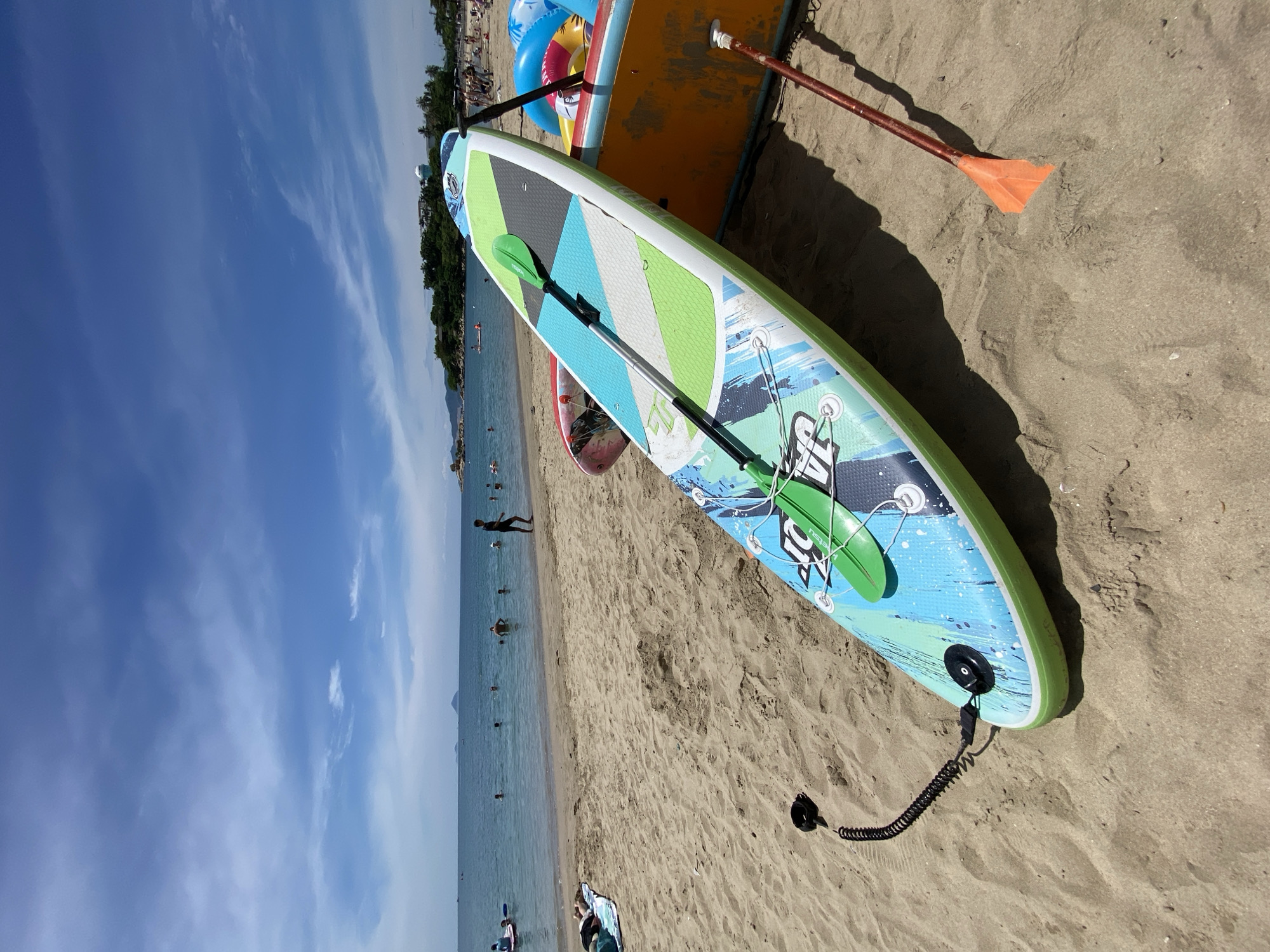 This image depicts a sunny day at a beach in Nha Trang, Vietnam, likely near a popular tourist area due to the presence of stand-up paddleboards and people enjoying the water. In the foreground, a blue and green patterned stand-up paddleboard with a green paddle rests on the sand, attached by a black coiled leash. To its right, an orange object, possibly another paddleboard or a floatation device, leans against it. Further back, the sandy beach is dotted with people, some relaxing on towels, while others are in the shallow ocean, swimming or paddling on surfboards and inflatable rings. The water is calm and a clear blue, with a distant coastline visible. The sky above is bright blue with scattered white clouds, indicative of good weather. The overall scene conveys a relaxed beach atmosphere common in Nha Trang during peak tourist seasons.