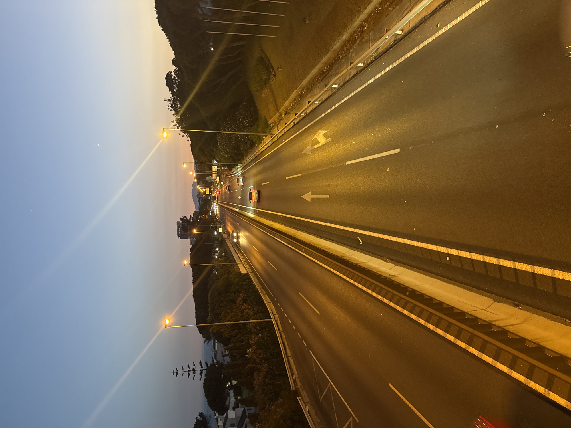 An elevated, forward-facing perspective captures a dual carriageway highway at dusk in Mijas, Spain. The highway is illuminated by streetlights, casting a warm orange glow on the asphalt. Lane markings are clearly visible, including solid and dashed white lines, and directional arrows painted on the road surface indicate traffic flow. Several cars are present on the highway, moving in both directions, with their headlights and taillights visible. The highway is bordered by a concrete barrier and guardrails. On either side of the highway, lush green trees and vegetation are visible. In the distance, buildings and residential structures can be seen nestled among the hills. The sky above is a gradient of deep blue and orange, indicating the transition from day to night. A faint crescent moon is visible in the upper right portion of the sky. The scene suggests a typical evening commute on a major road in a semi-urban or suburban area.
