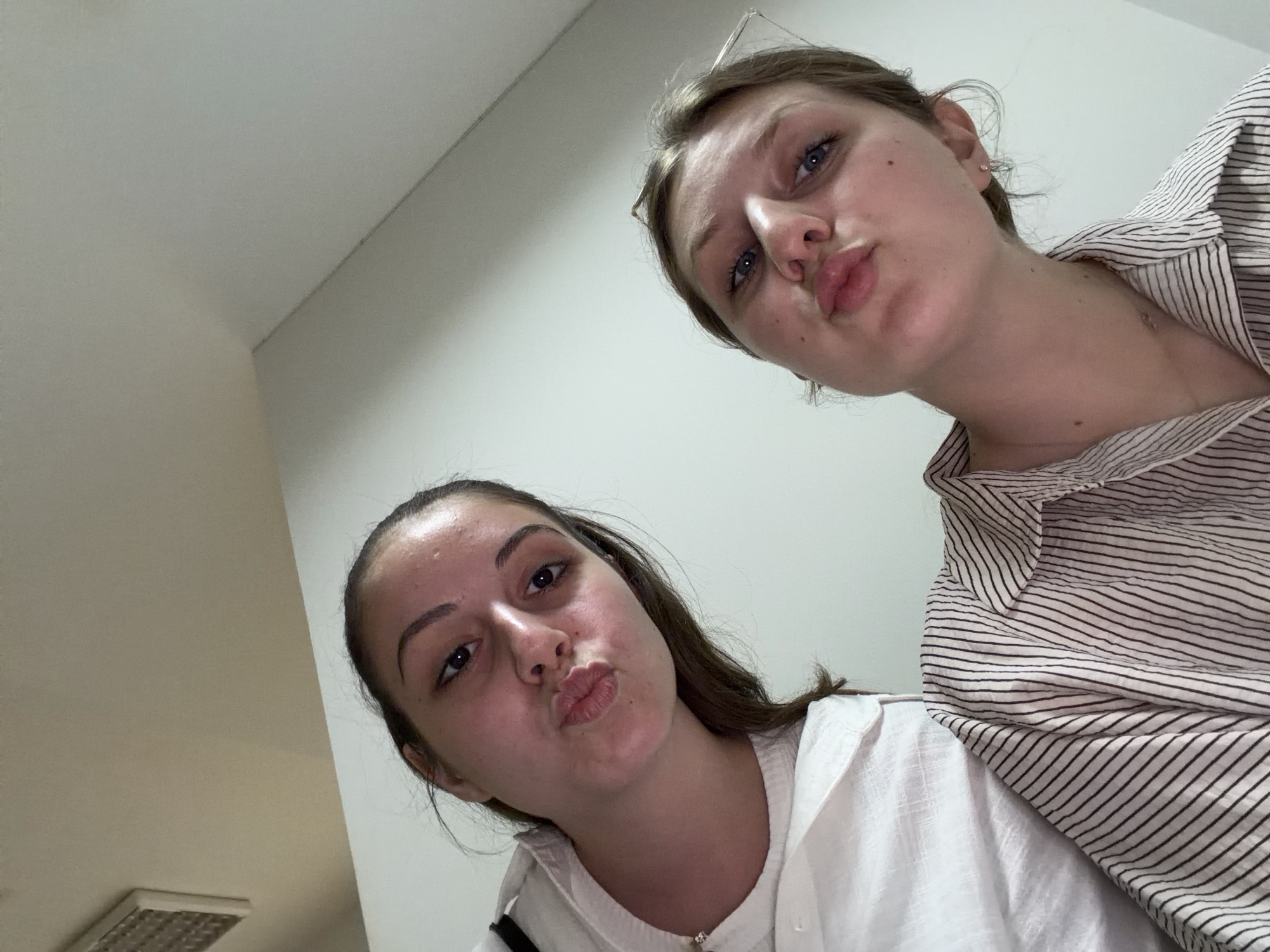 This is a selfie taken by two young women in an indoor environment. The woman on the left has dark brown hair and is wearing a white top. She is puckering her lips and looking at the camera. The woman on the right has blonde hair and is wearing a striped shirt. She is also puckering her lips and looking at the camera. The background appears to be a wall, possibly in a room. There is a vent visible on the ceiling in the upper left corner of the image. The lighting suggests it is daytime, possibly indoors with artificial lighting. No specific location within Burgas, Bulgaria can be identified from the image.