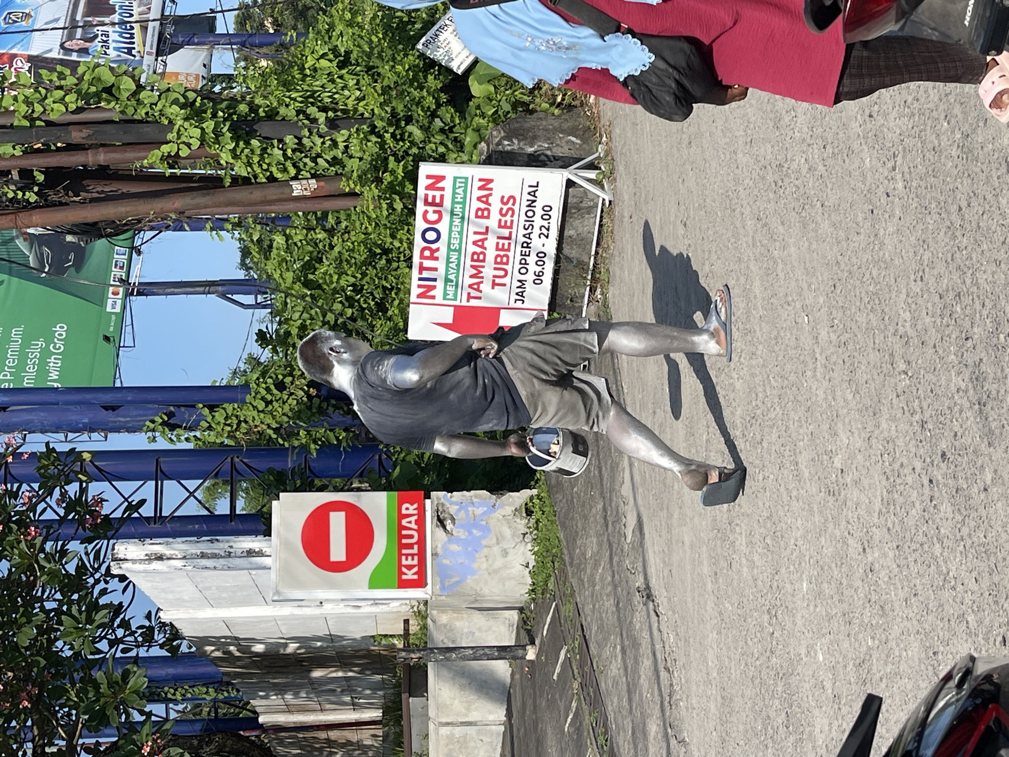 A person covered in silver paint walks on a paved street in Kuta, Indonesia. They are wearing shorts and flip-flops, and carrying a white bucket with a handle, also covered in silver paint. The person's body is mostly covered in silver, with some areas showing darker skin tones through the paint. In the background, there is a white sign with a red circle and a horizontal bar, indicating "KELUAR" (exit). Further in the background, there is a white sign advertising "NITROGEN TAMBAL BAN TUBELESS" with operating hours from 06:00 to 22:00. A person wearing a blue hijab and a red shirt is visible on the right side of the image. The environment appears to be an urban street with vegetation and utility poles. The scene is illuminated by bright sunlight, casting shadows on the ground.