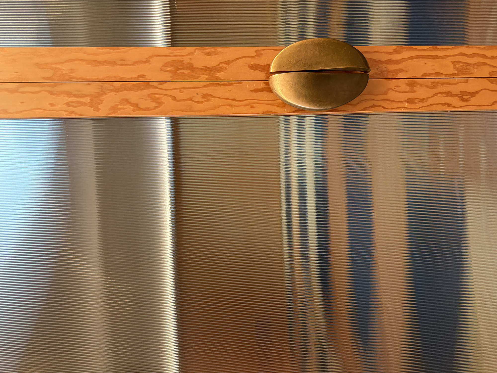 This close-up image presents an abstract view of a textured surface, likely a door or wall, in Los Angeles, United States of America.
The primary subjects are a central horizontal wooden plank and a metallic decorative or functional element. The wooden plank is a light orange-brown color, displaying a prominent, swirling wood grain pattern. It appears to be composed of two narrower planks joined horizontally, indicated by a faint dark line running its length.
Positioned on the right side of this wooden band, partially overlapping it, is a circular, split metallic object. This object, likely a handle or an ornate pull, possesses a warm brass or bronze hue with some areas showing a slight patina, and is divided horizontally into two distinct halves.
Above and below the wooden plank, the surface features a consistent pattern of very fine, horizontal ridges or grooves. This corrugated or fluted texture is highly reflective, casting elongated vertical streaks of varying light and shadow across its surface. The reflections range in color from cool, bright blues and silvers on the left to darker, warmer browns and blacks on the right, suggesting the influence of ambient lighting and possibly other objects within the immediate environment.
No activity or event is taking place, as the image captures a static architectural or furniture detail. There are no discernible time of day or weather cues, and the lighting appears to be indoor or controlled. No text is visible in the image. The reflections provide the only subtle hint of the surrounding space.