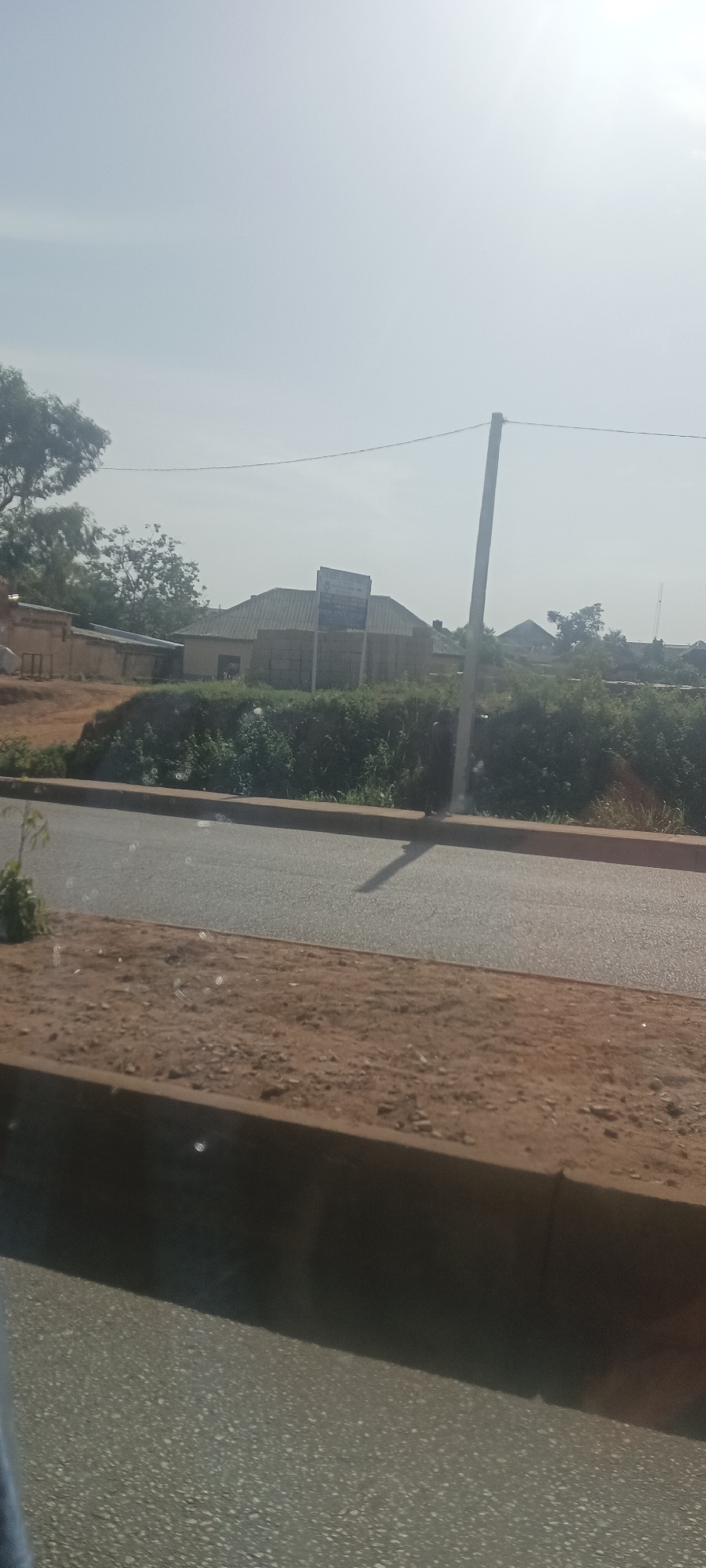 The image shows a street view in Bukuru, Nigeria, under a bright, hazy sky. A gray asphalt road runs horizontally across the midground, bordered by a dirt shoulder and a concrete curb. Beyond the road, a dense strip of green vegetation obscures the lower portions of buildings, some of which appear to be single-story structures with metal roofs. A utility pole stands near the roadside, casting a shadow on the pavement. A sign is visible atop one of the buildings behind the vegetation. The foreground shows a portion of a curb and road, suggesting the image was taken from a vehicle.